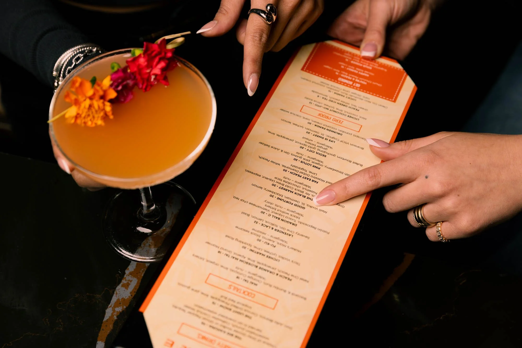 Close-up of person holding a cocktail glass with orange beverage and flower garnish, pointing to a long orange-colored menu on a black surface.