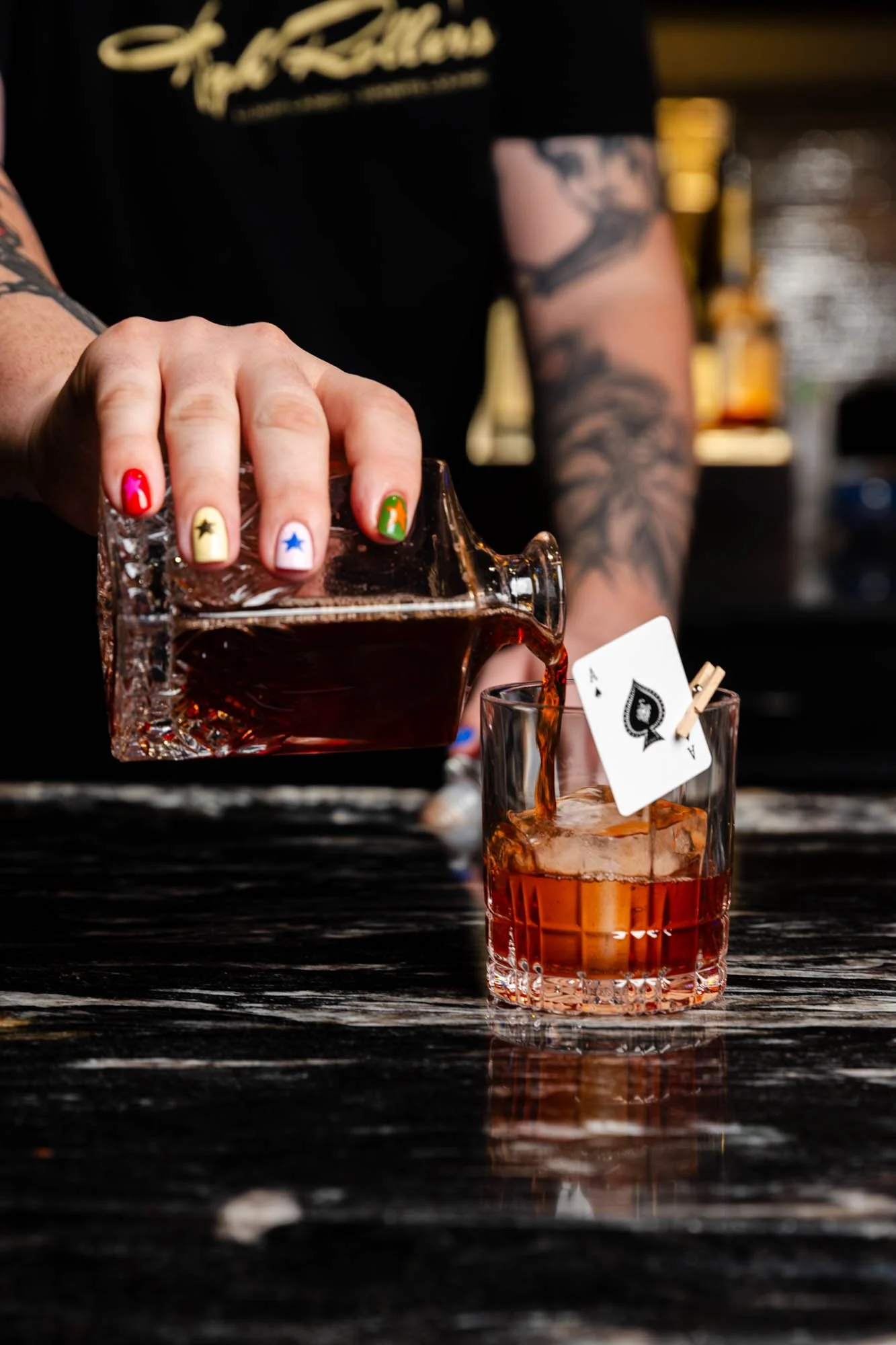 Person pouring a dark alcohol from a glass decanter into a glass with ice and a playing card, 'Ace of Spades', clipped to the glass, in a bar setting.