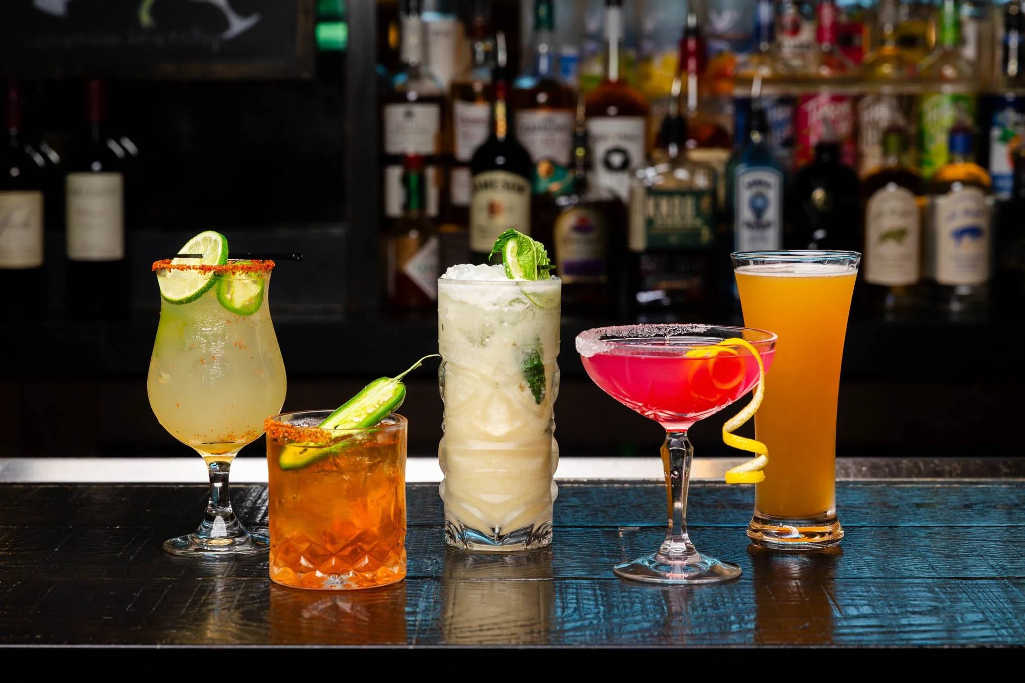 Six colorful cocktails on a dark bar counter with a blurred background of liquor bottles. food and beverage photography. 