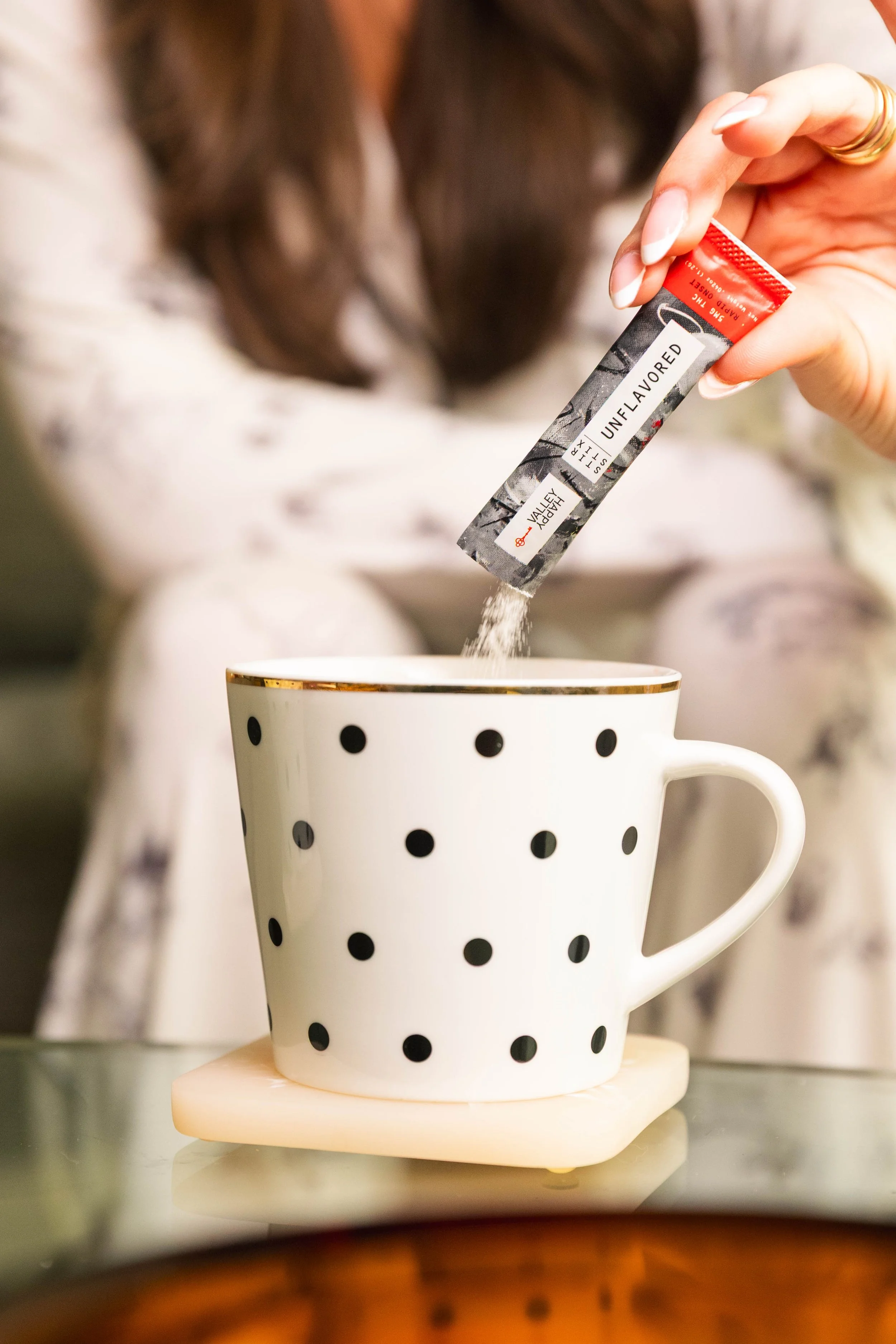A woman is pouring unflavored salt from a red and white packet into a polka dot mug, which is sitting on a coaster.