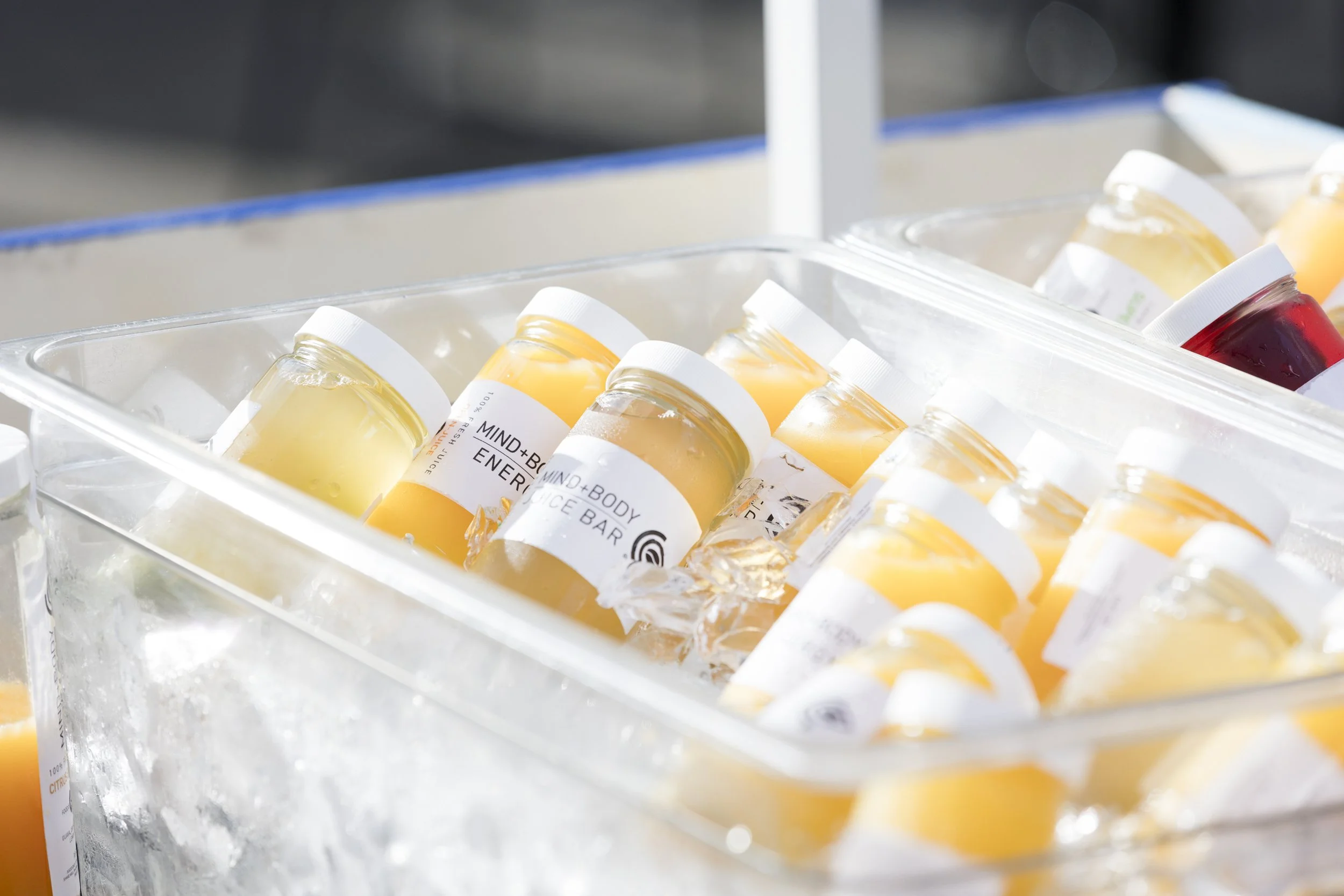 Several small bottles filled with yellow, orange, and red liquid, labeled with health or energy-related names, are stored in a clear ice-filled container. event photography.
