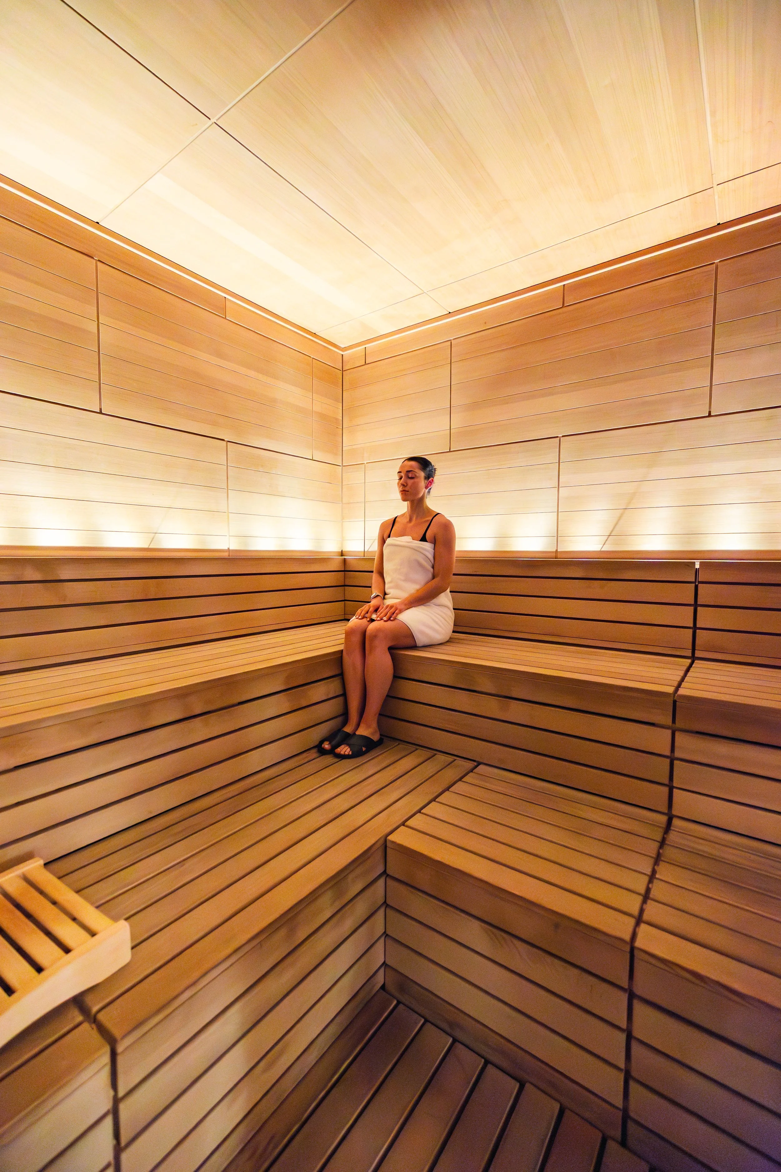 A woman sitting in a wooden sauna, wrapped in a towel with her eyes closed and hands resting on her lap, surrounded by warm lighting and wooden walls. lifestyle and hospitality photo. 
