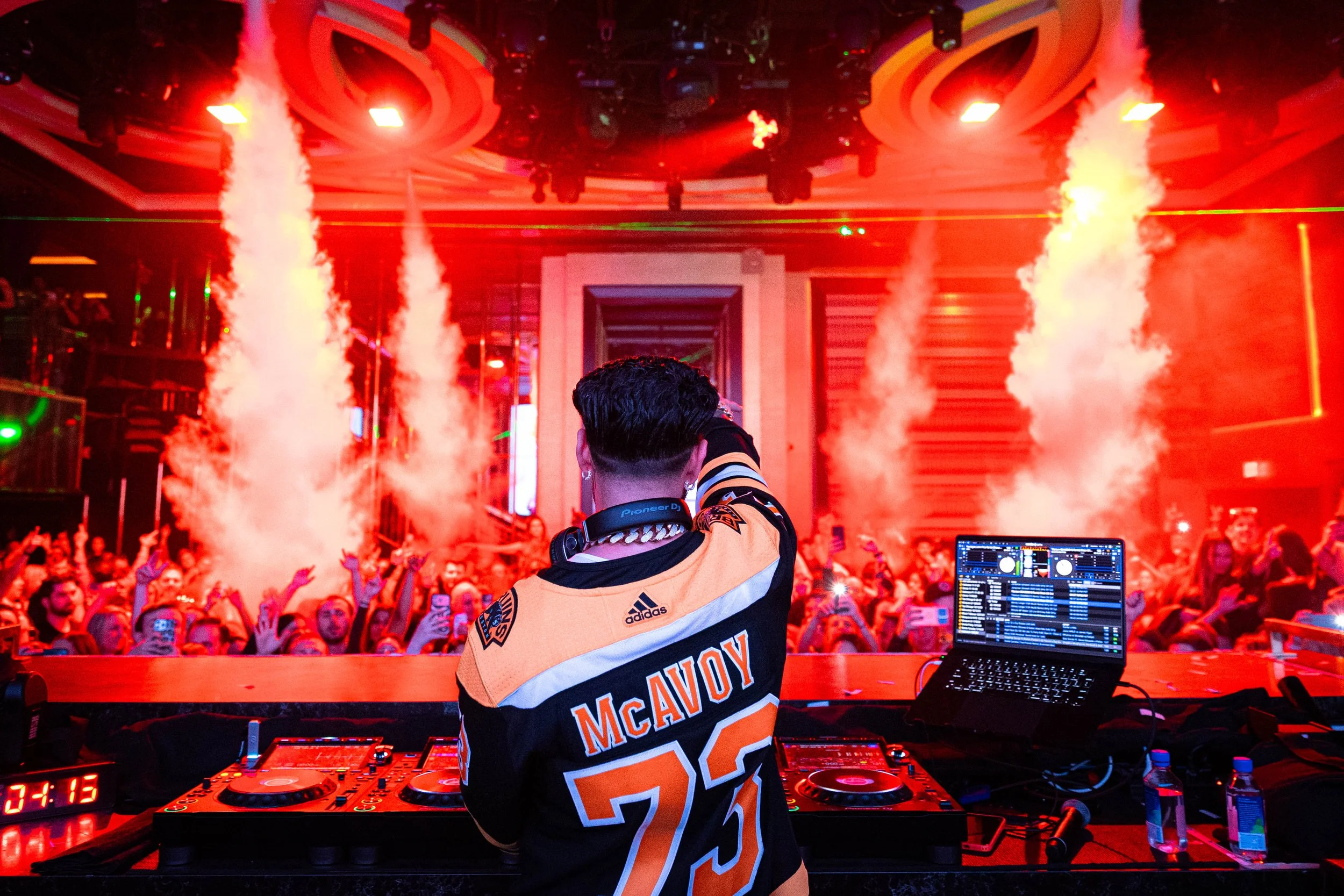 DJ Pauly D is performing at a lively indoor concert, with red and green laser lights and smoke effects. The crowd is visible in the background, enjoying the music. 