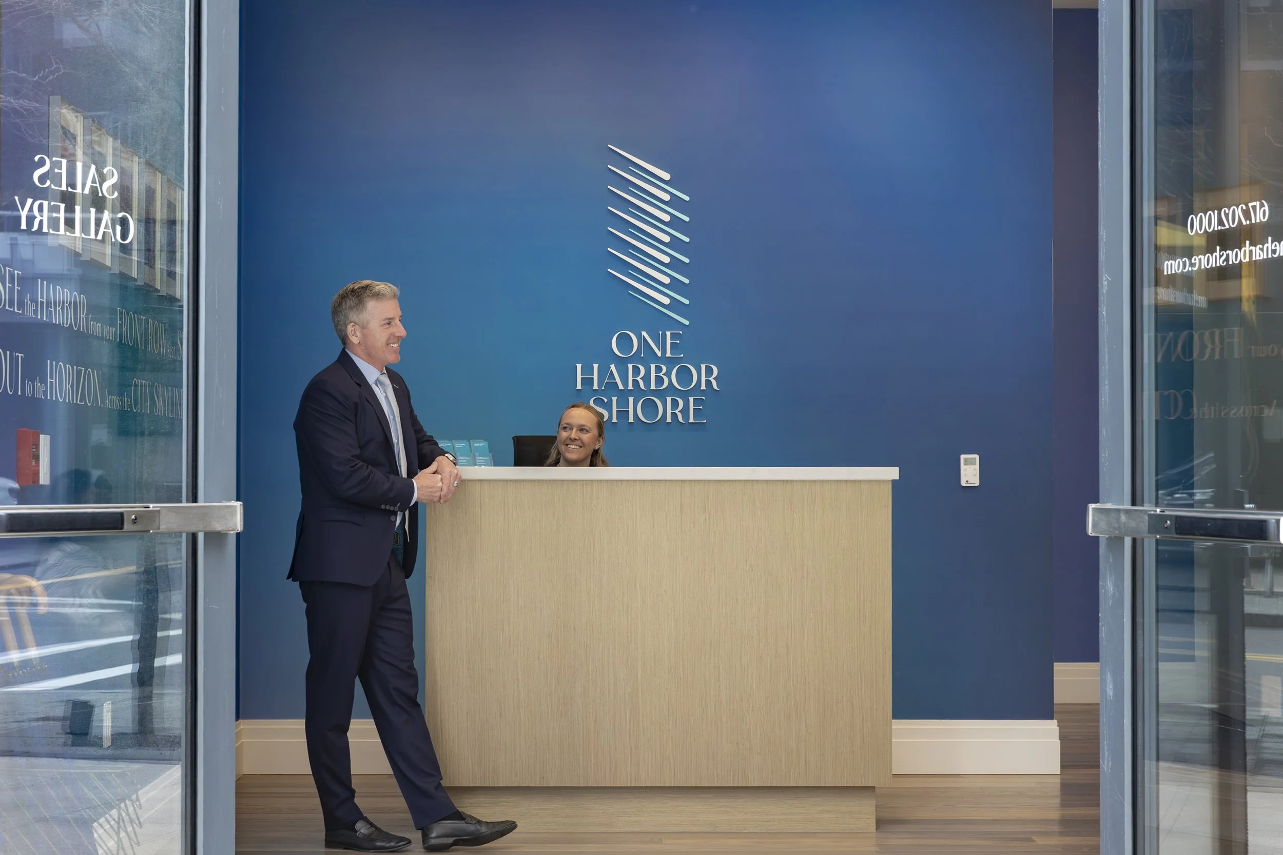 A man and a woman smiling and talking at a reception desk inside a building with a sign that reads "One Harbor Shore." The man is dressed in a suit and the woman is behind the desk.