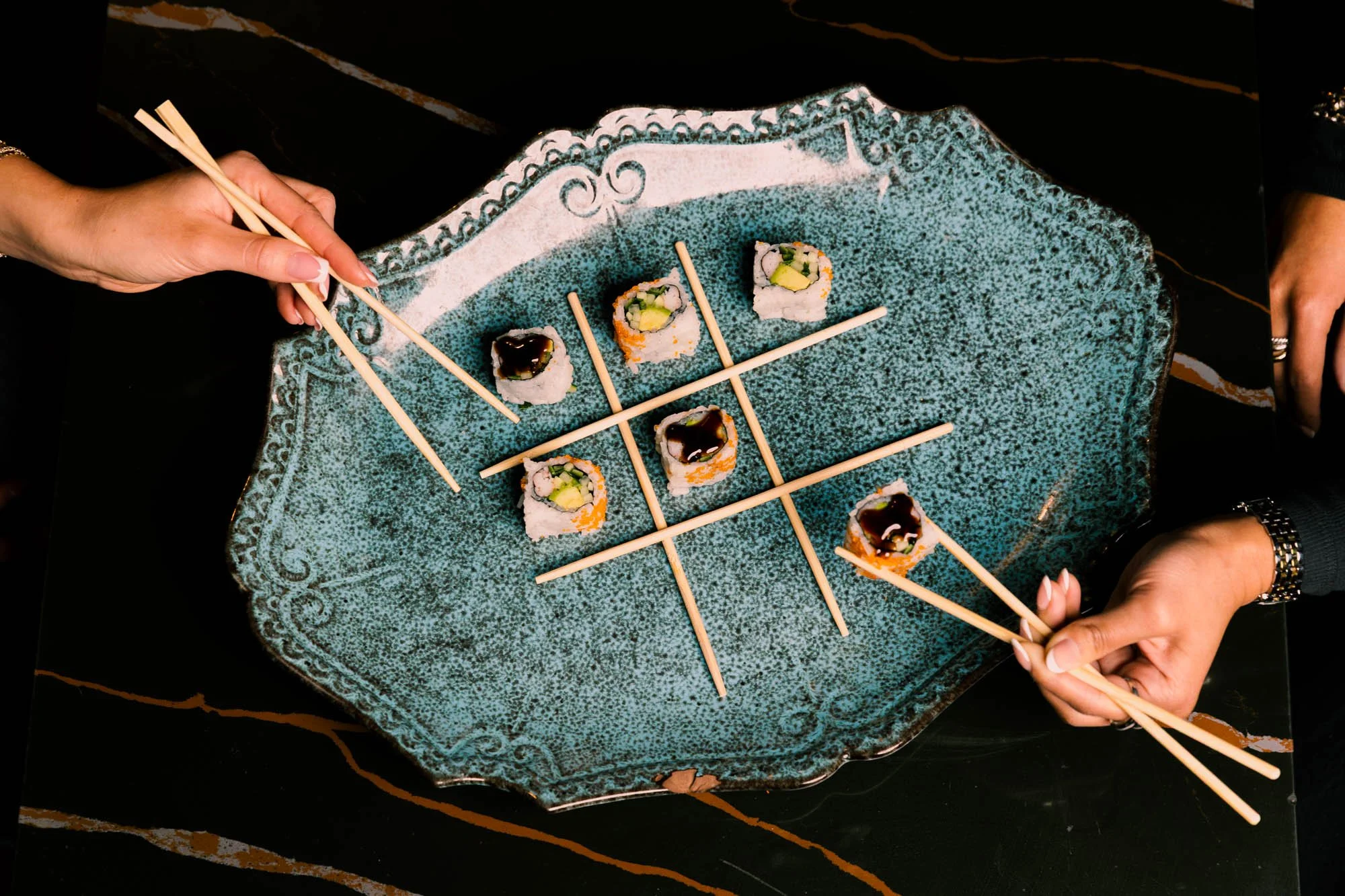 A dark green textured platter holds assorted sushi rolls with different fillings and toppings, with a person using chopsticks to pick up a piece. Hands are visible on either side of the platter.