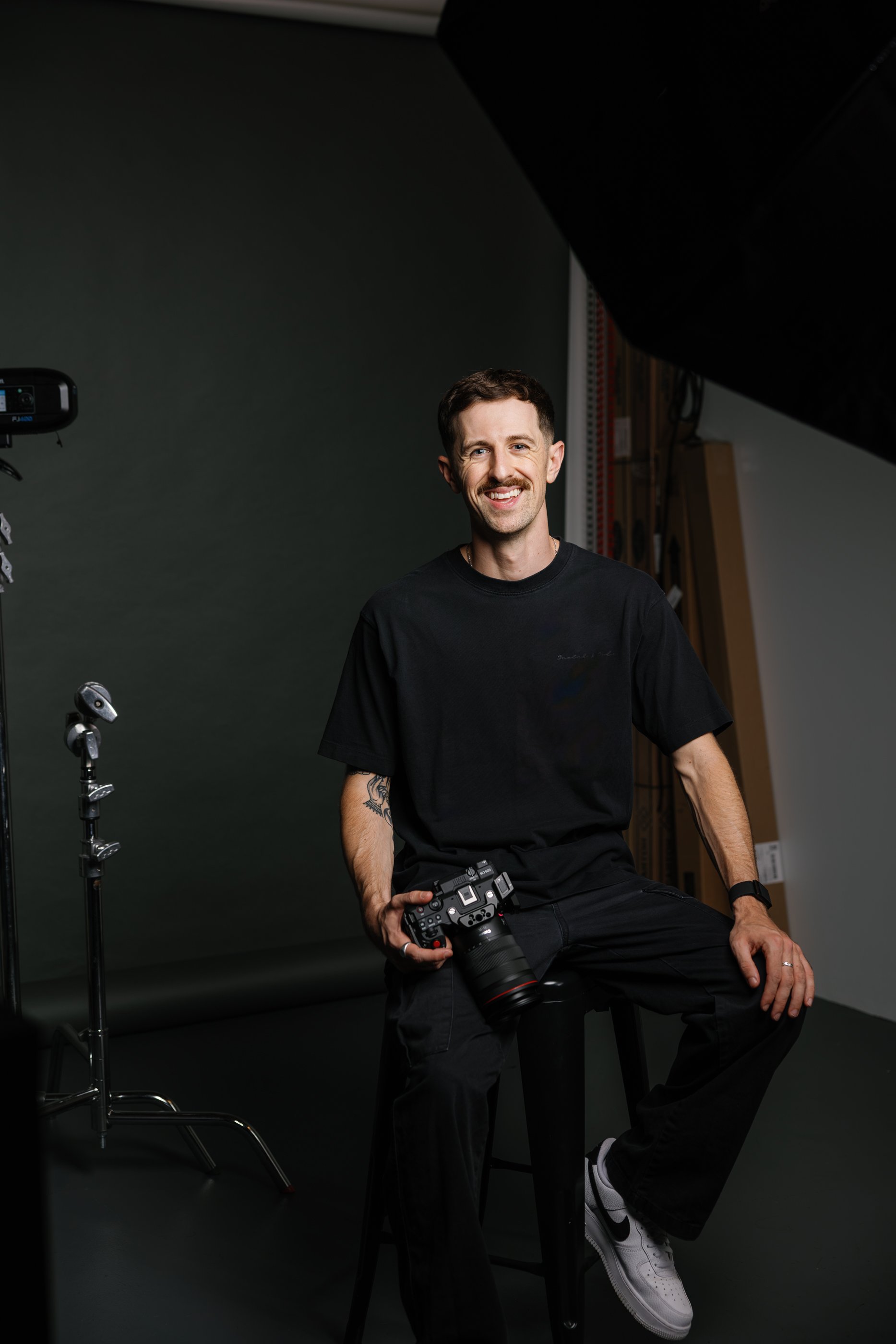 A smiling man with short dark hair, a mustache, and tattoos on his left arm, holding a professional camera, sitting on a stool in a photography studio with dark walls, professional lighting, and equipment around him.