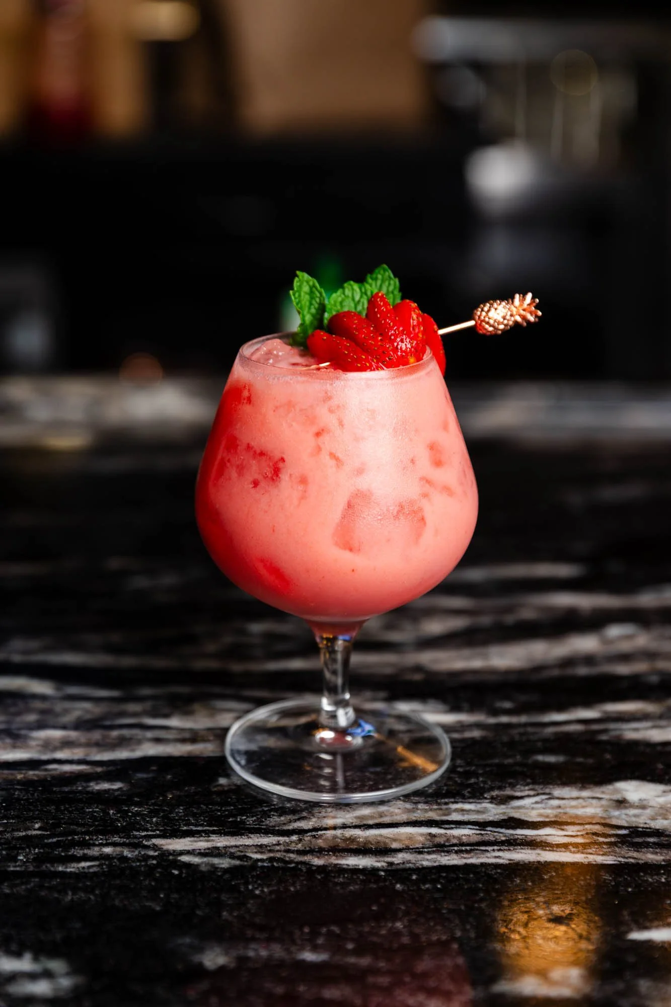 A pink strawberry daiquiri cocktail garnished with sliced strawberries, fresh mint leaves, and a decorative stir stick, served in a stemmed glass on a dark marble surface. food and beverage photography.