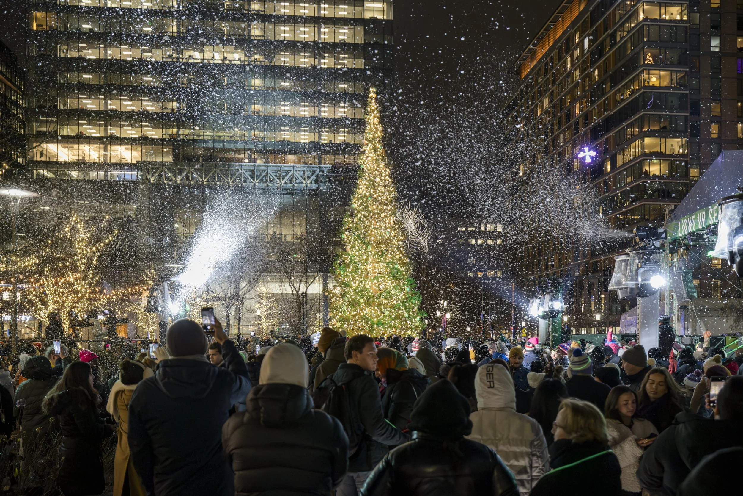 Group of people gathered outdoors at night for a Christmas celebration, with a decorated Christmas tree, artificial snow, and city buildings illuminated in the background. event photography
. 