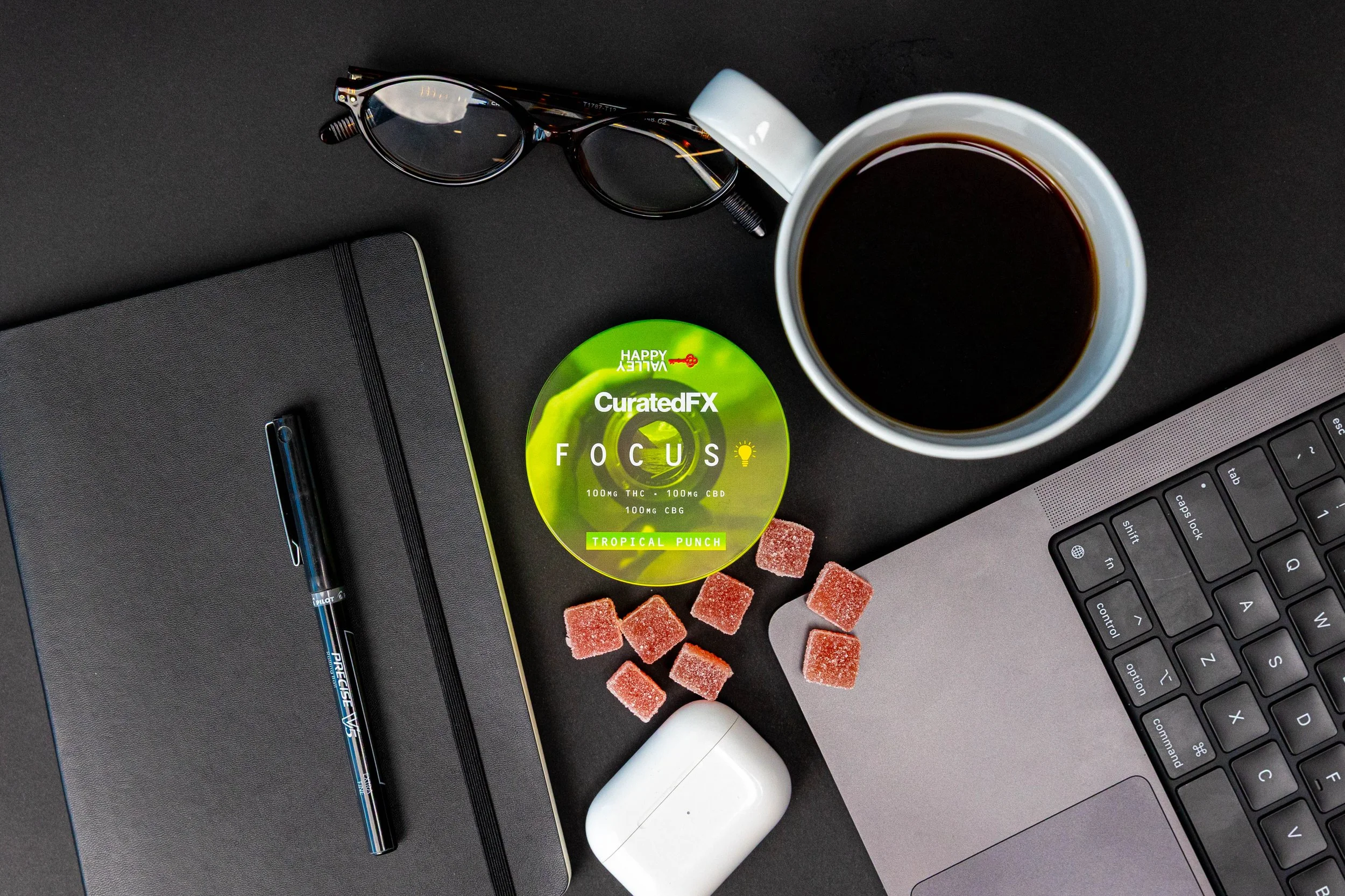 A workspace with a notebook, pen, glasses, a cup of black coffee, a laptop, a cannabis focus product labeled 'CuratedFX,' and sugar cubes on a black surface.