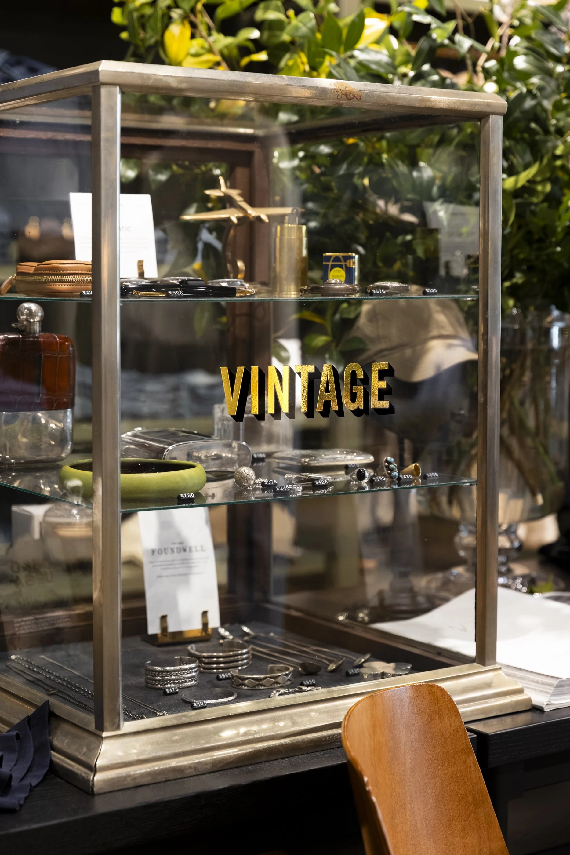 Display case with vintage jewelry and accessories, including rings, bracelets, and necklaces, in a store setting with greenery background and a wooden chair in the foreground.
