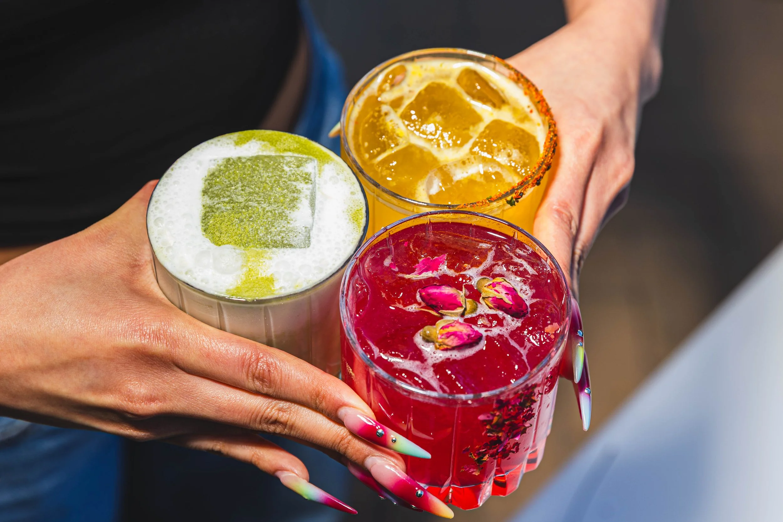 A person is holding three colorful drinks with ice cubes and flower garnishes, using long, decorated nails. food and beverage photography.