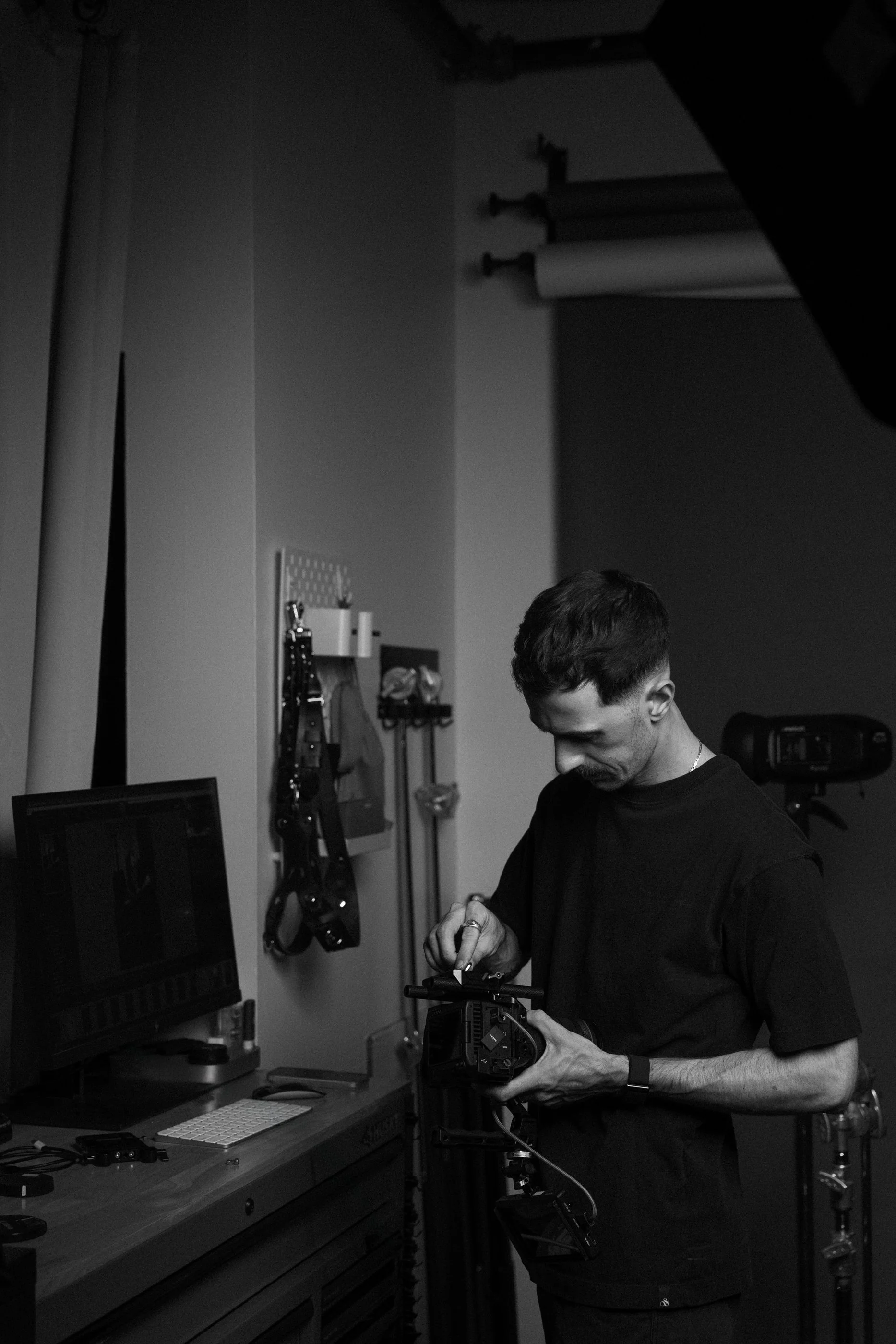 A man working in a photography or videography studio, adjusting camera equipment at a workstation with monitors, surrounded by various tools and gear hanging on the wall.