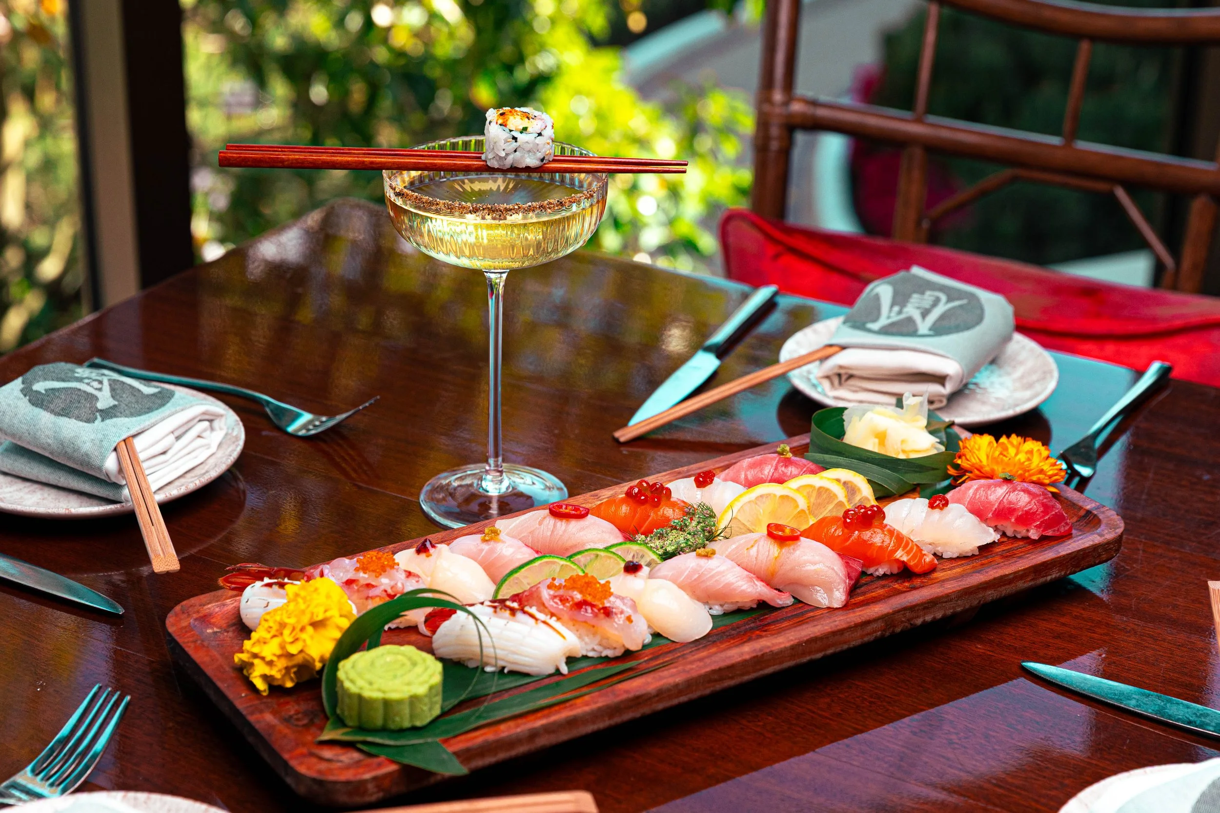 Sushi platter with various types of sushi including nigiri and sashimi, garnished with flowers and citrus slices, set on a wooden table with utensils and napkins, in an outdoor setting. Jack MacMullin, Boston, Seaport, Photographer, Videographer, Eve