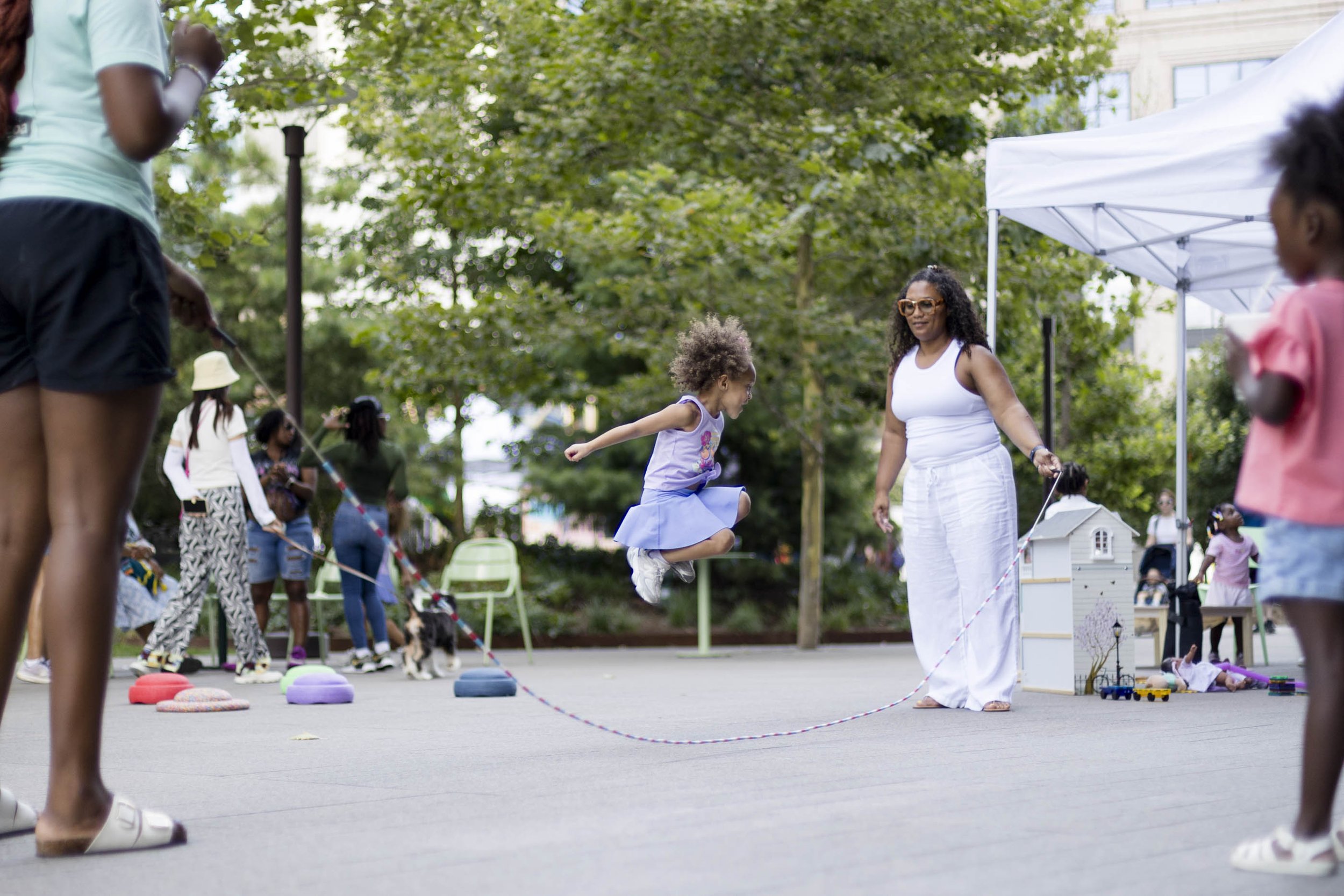 Children and adults playing and walking at an outdoor park or community event with trees, tents, and various toys and equipment set up. event photography. 