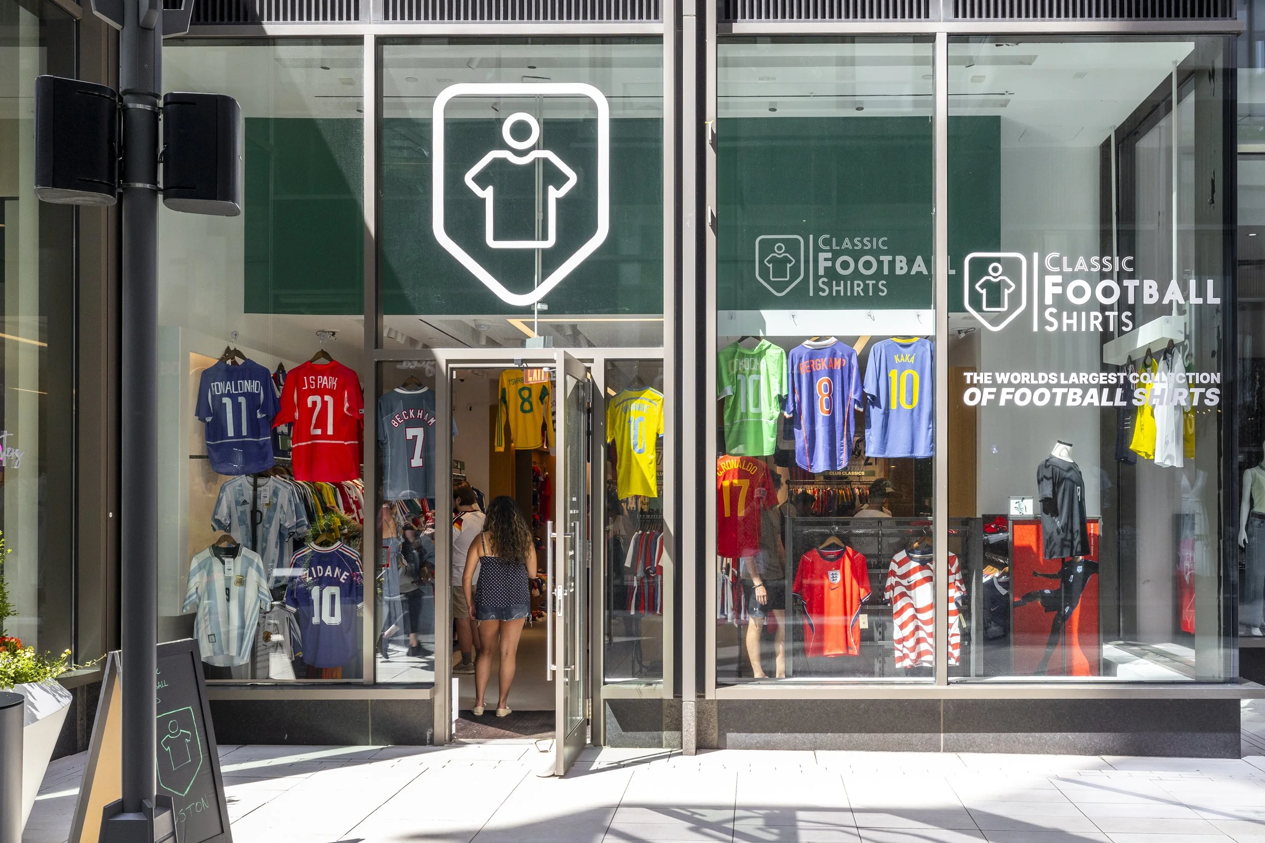 Storefront window display of a shop selling football shirts, featuring various jerseys of different teams and players, with a large logo and text advertising 'Classic Football Shirts'.