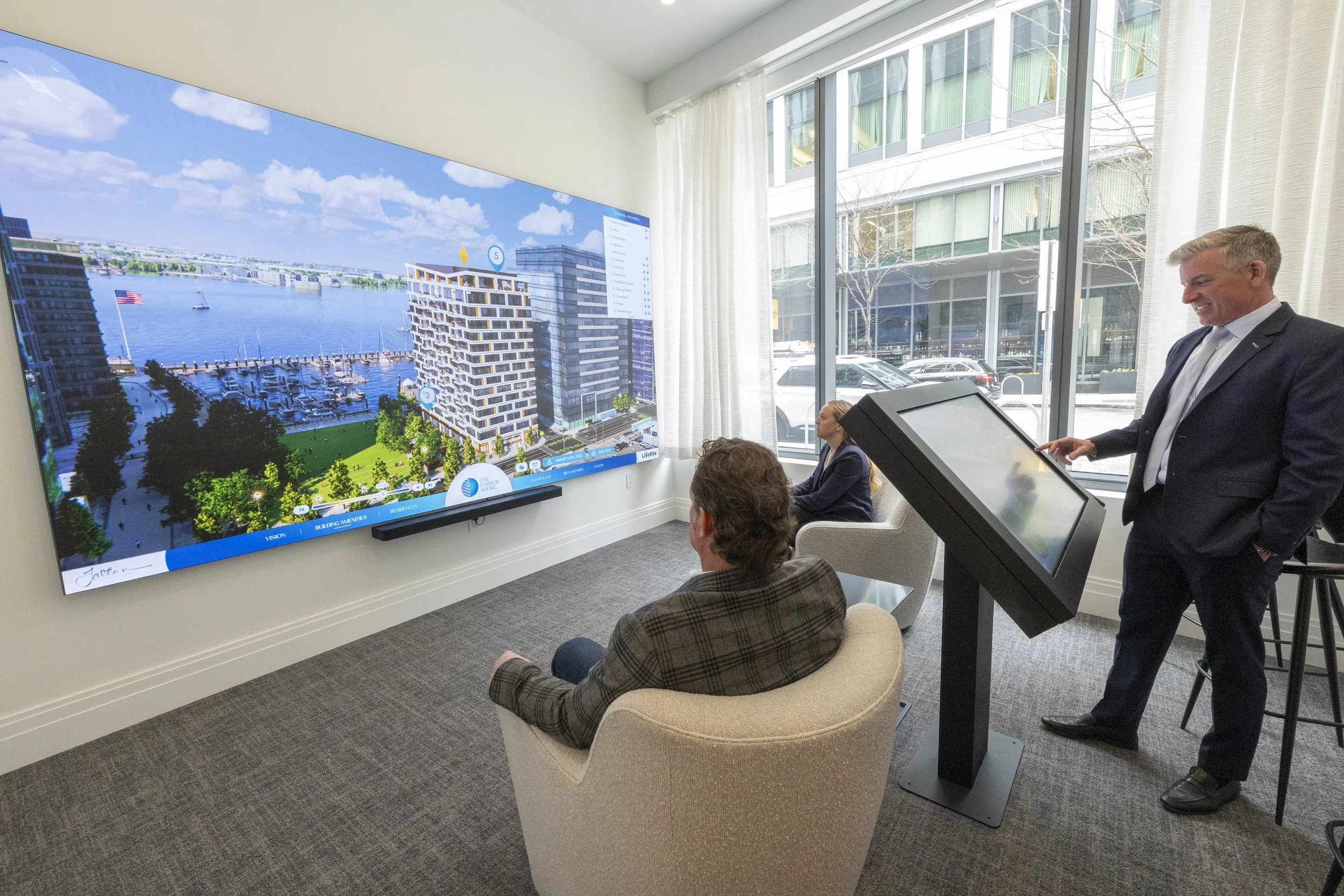 People attending a presentation in a conference room with a large screen displaying a building plan and waterfront scene; one man in a suit is standing near an interactive kiosk, smiling.