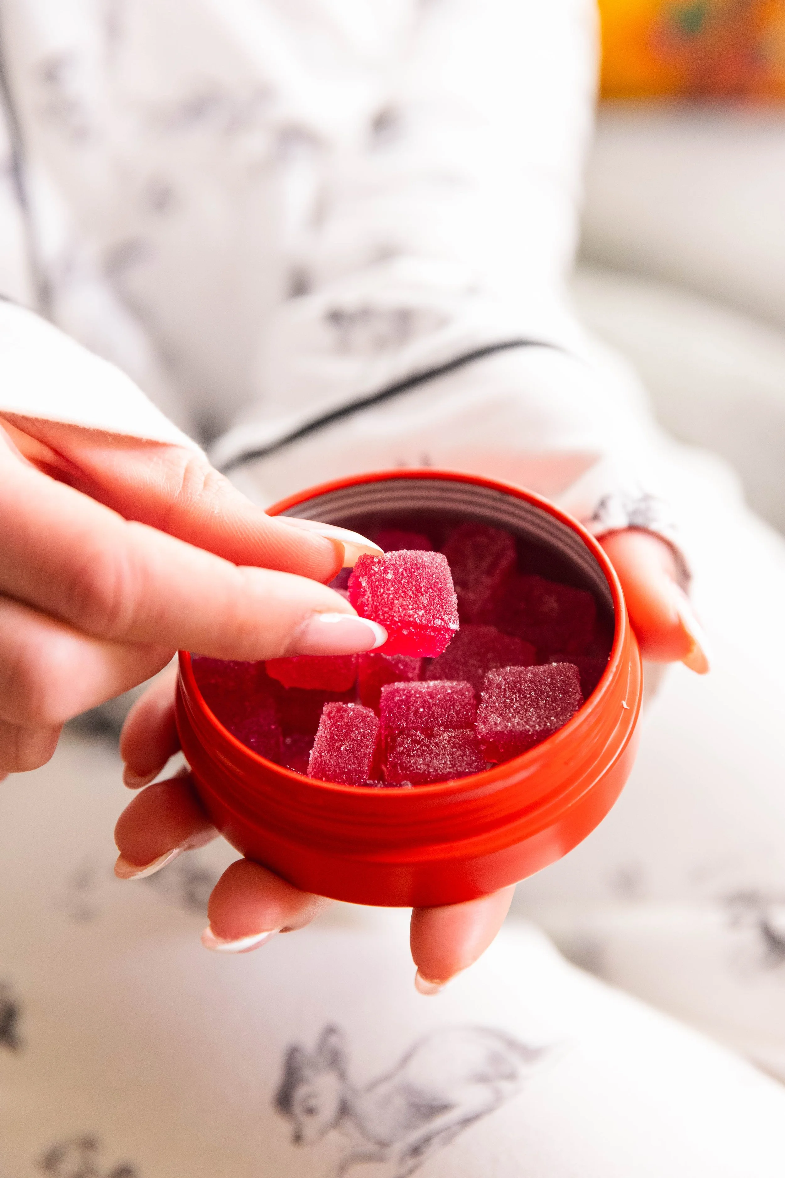 Person with well-manicured nails holding an orange container filled with red gummy bears, with one gummy bear being taken out.