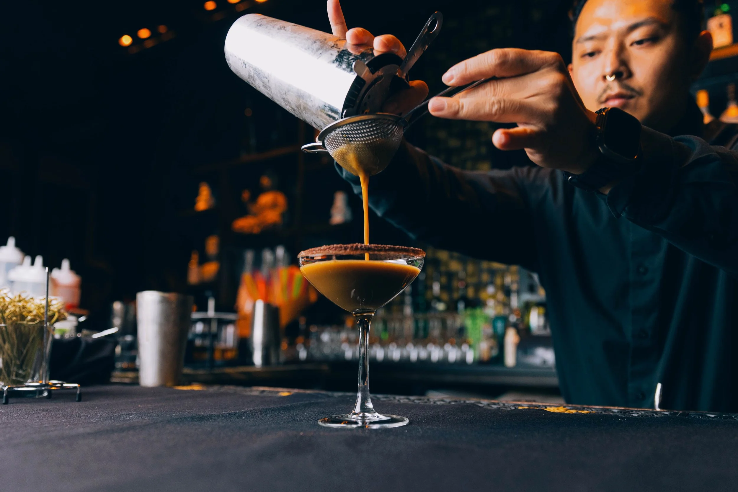 A bartender pours a cocktail through a strainer into a martini glass, with various bar tools and bottles in the background.