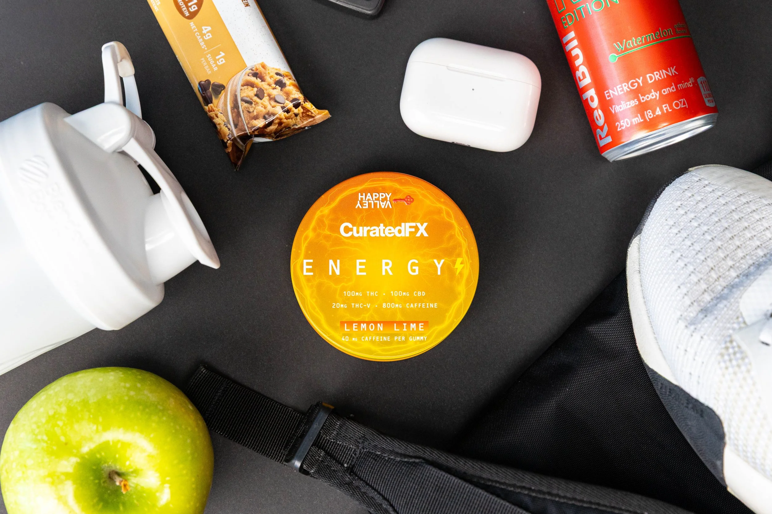 A black surface with various items including a white wireless earbud case, a yellow energy gummy container labeled CuratedFX Energy, a red Raspberry edition energy drink can, a white sneaker, a green apple, and a black bag strap.