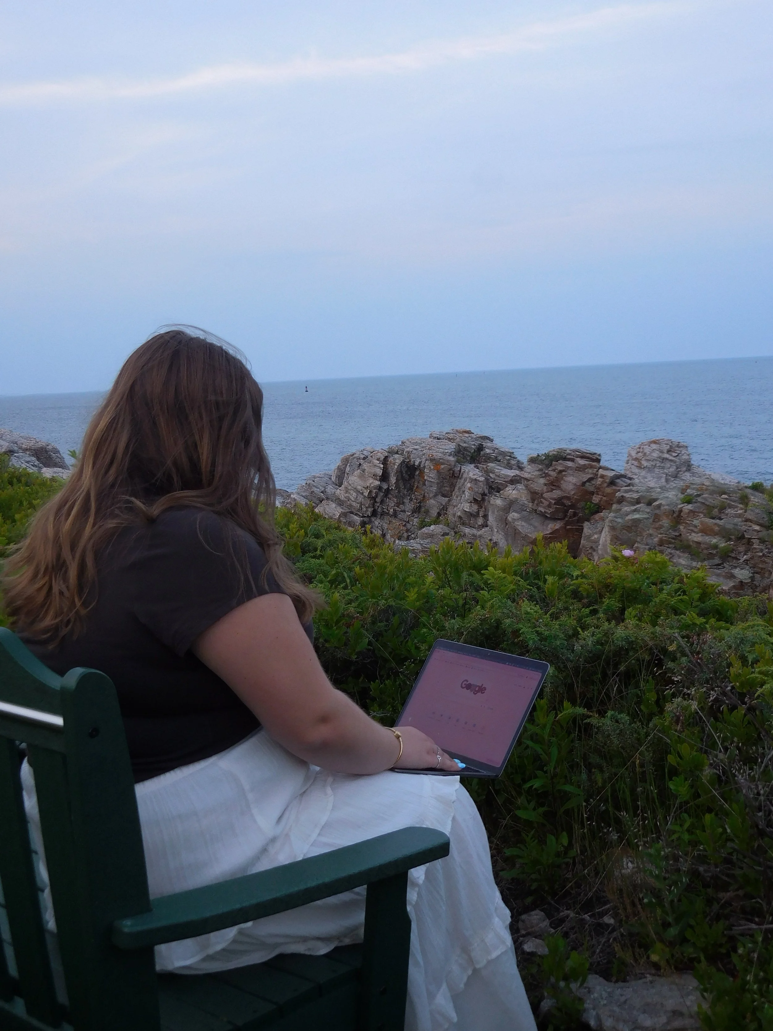 A woman with long brown hair sitting on a green outdoor chair near rocky coastal terrain, using a laptop with the Google homepage visible on the screen, with the ocean and sky in the background.