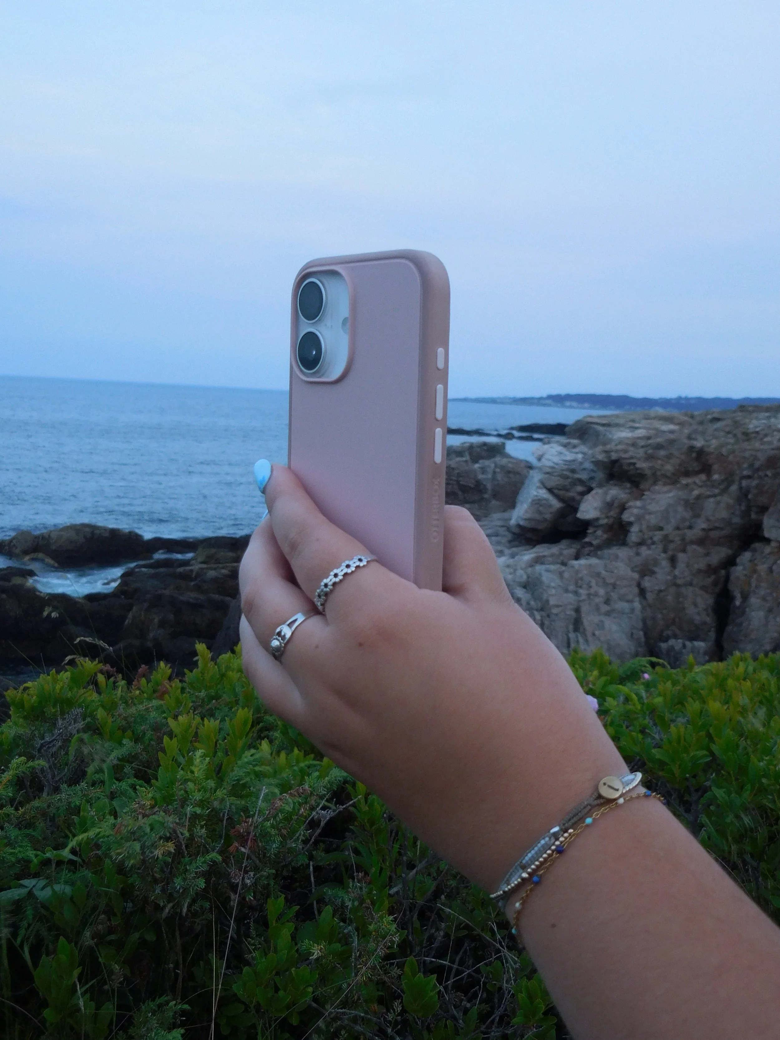 A person's hand holding a pink smartphone taking a photo of the ocean at sunset with rocks and green bushes in the foreground.