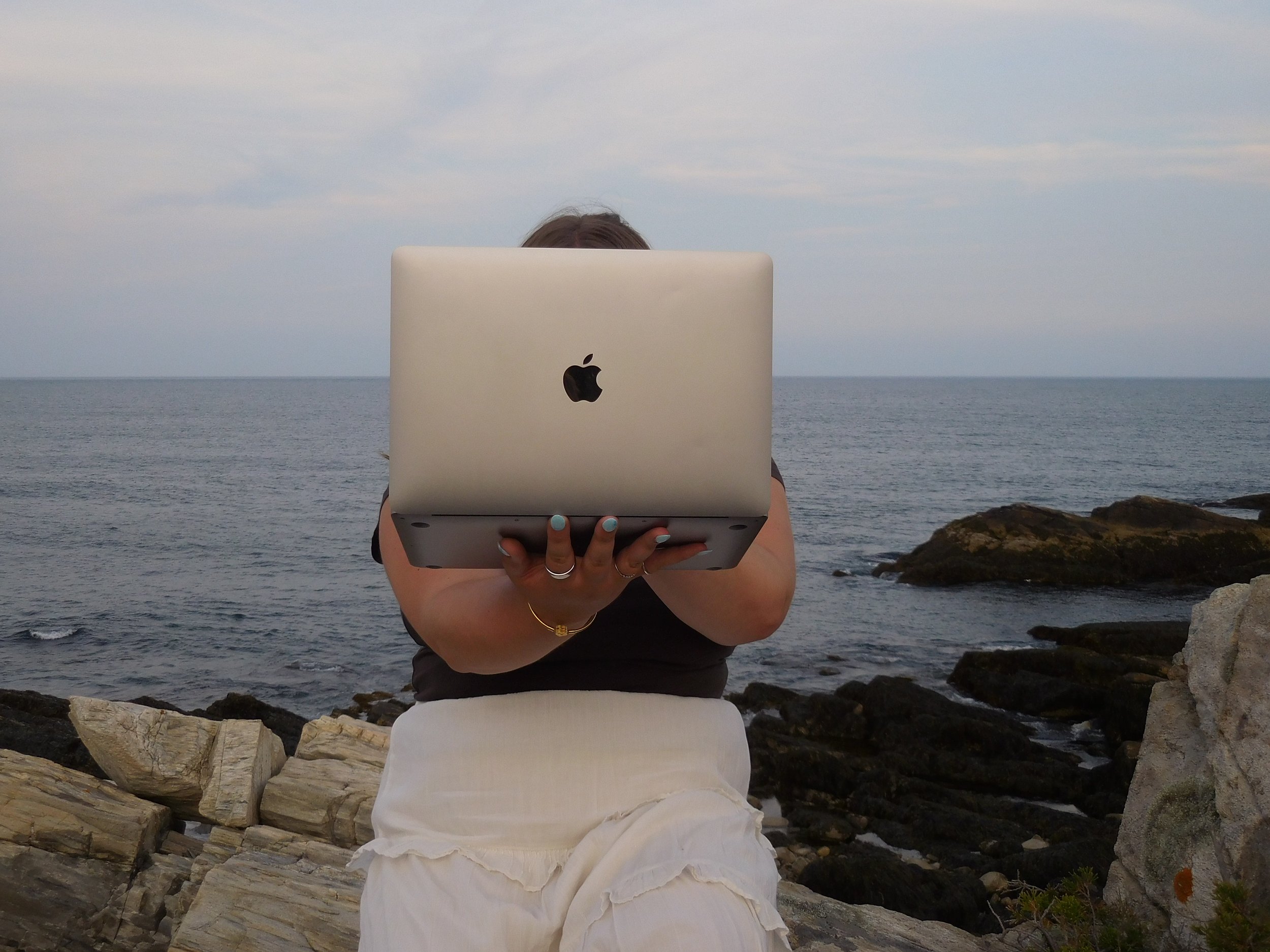 Person sitting on rocks by the ocean, holding an open MacBook with the Apple logo visible, during cloudy weather.