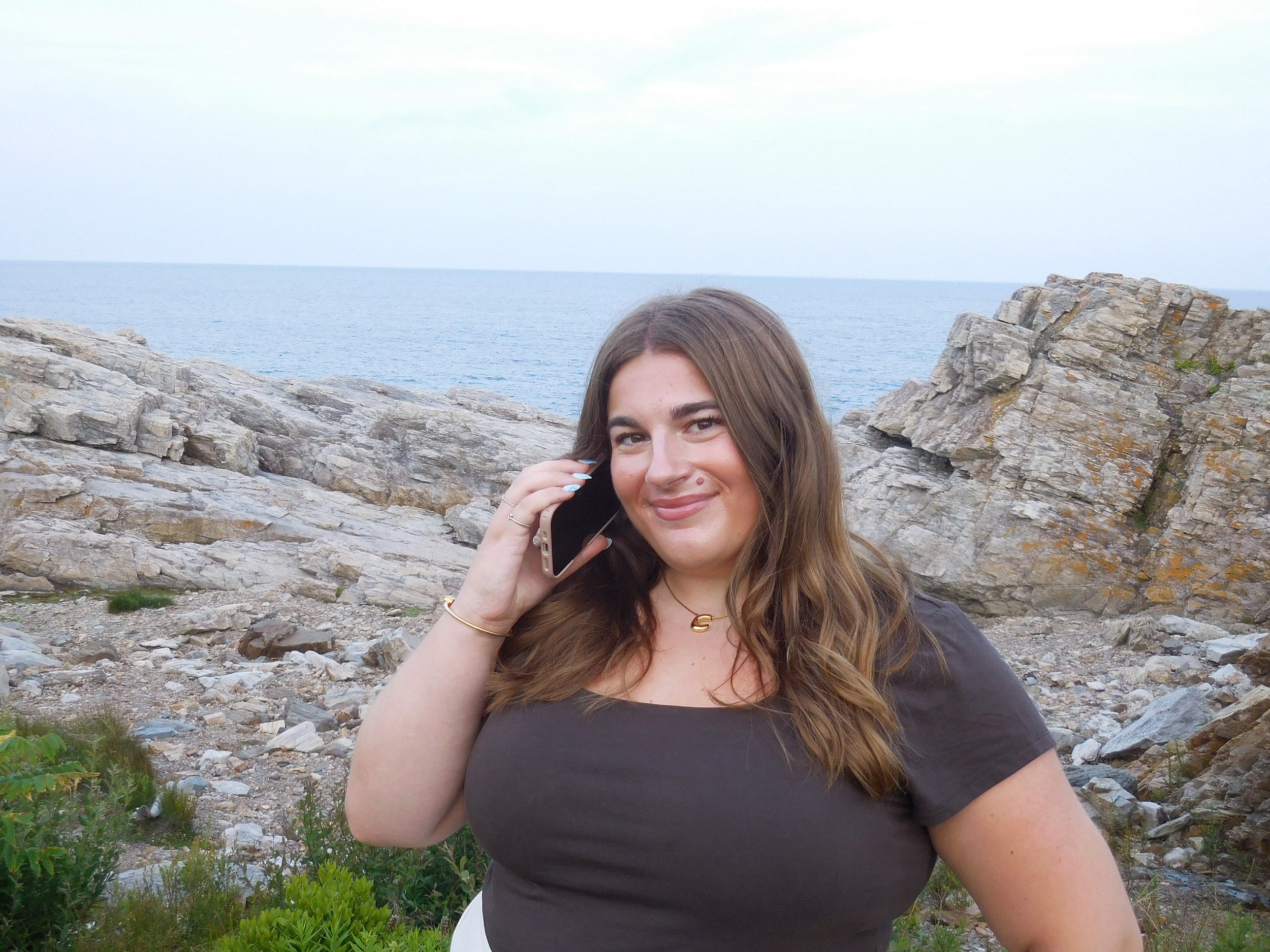 A young woman with long brown hair, smiling and talking on a cellphone, stands outdoors on a rocky coastline with the ocean and cloudy sky in the background.