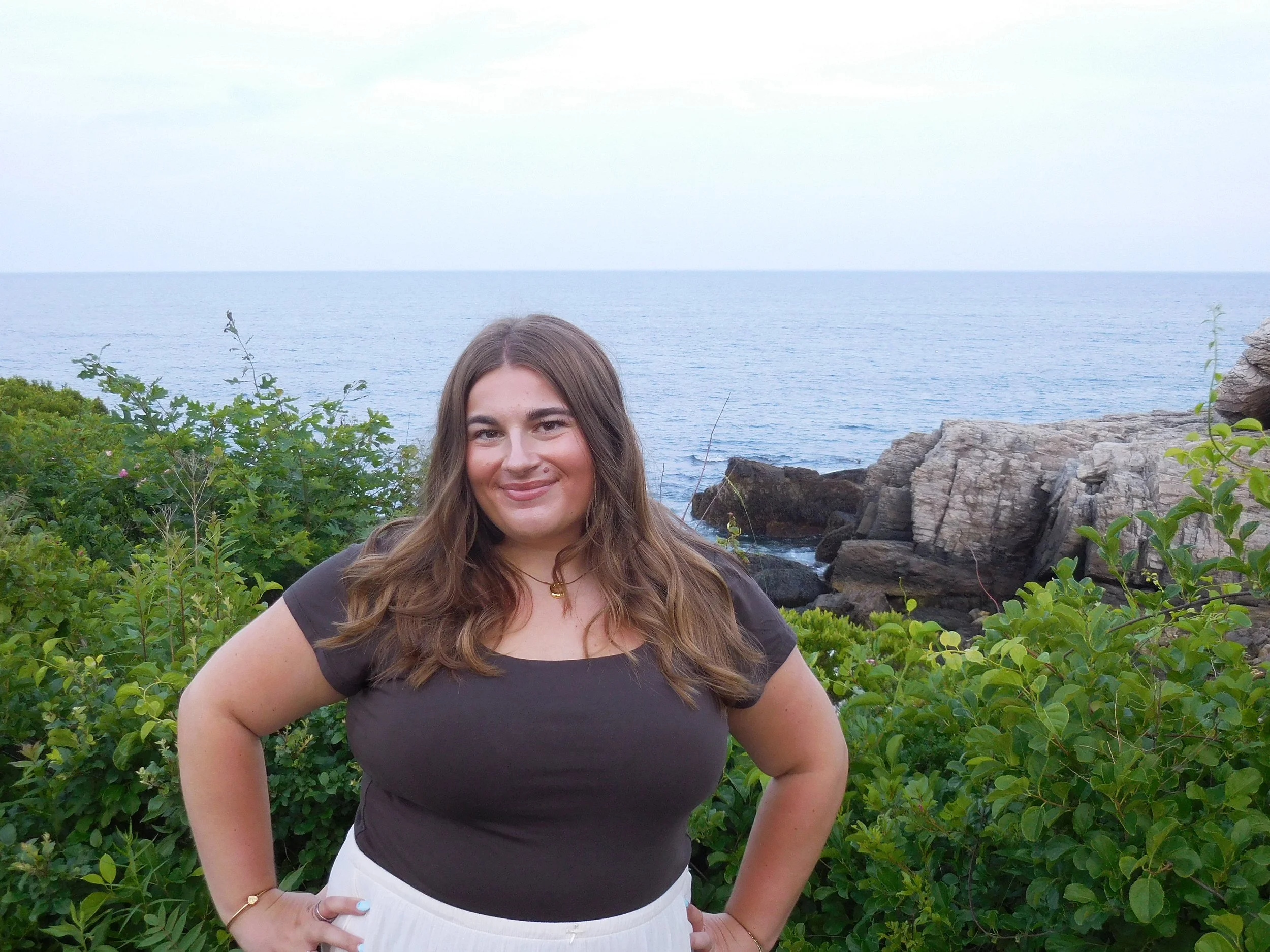 A woman standing outdoors with her hand on her hip, smiling, in front of green bushes, with rocks and water in the background.