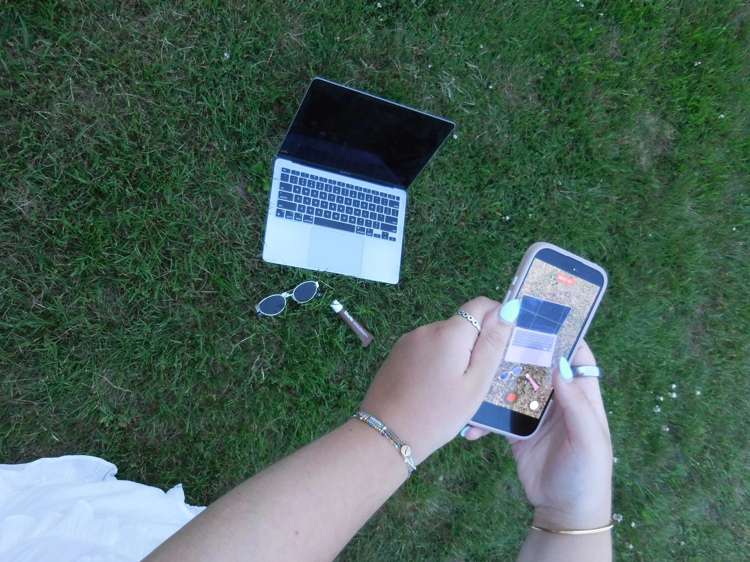 Person holding a smartphone while sitting on green grass, with a laptop, sunglasses, and a pen nearby.