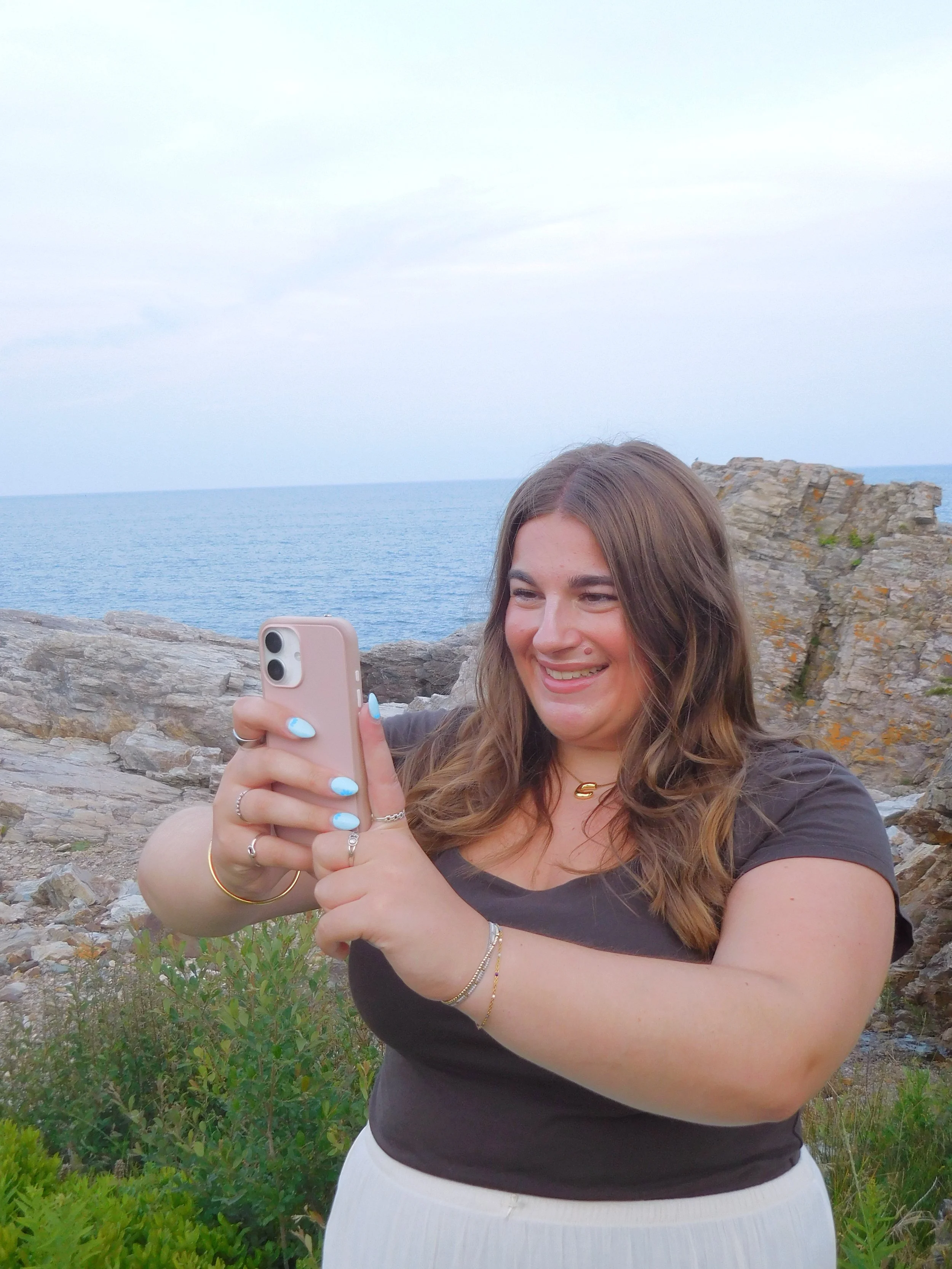 A woman with brown hair, wearing a black shirt and white skirt, taking a selfie with a smartphone on a rocky shoreline with the ocean and cloudy sky in the background.