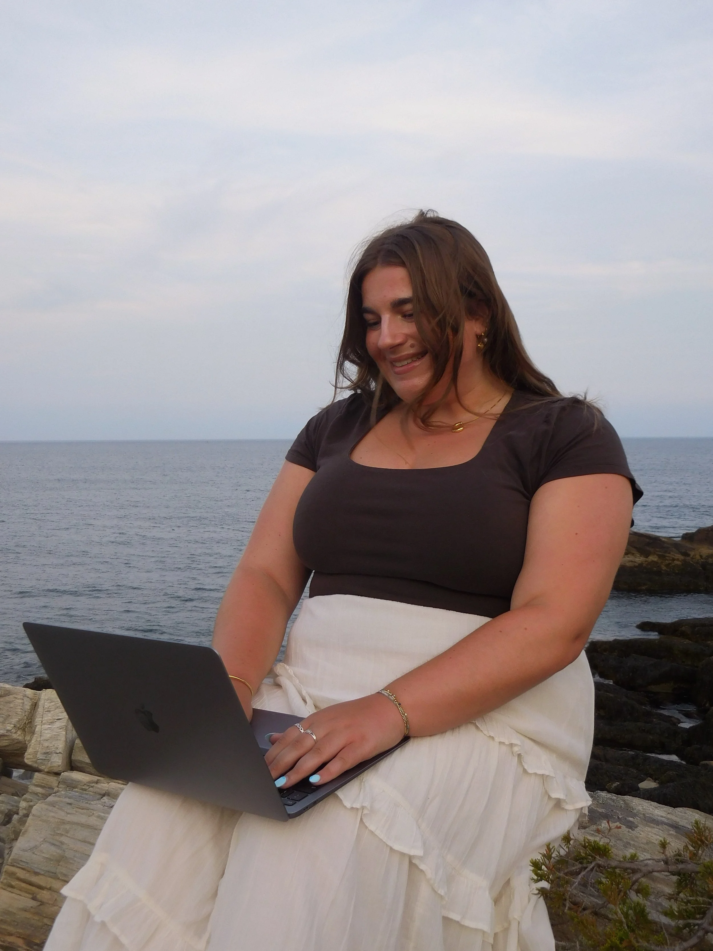 A woman sitting outdoors in front of the ocean, using a laptop, with rocks and some greenery nearby.