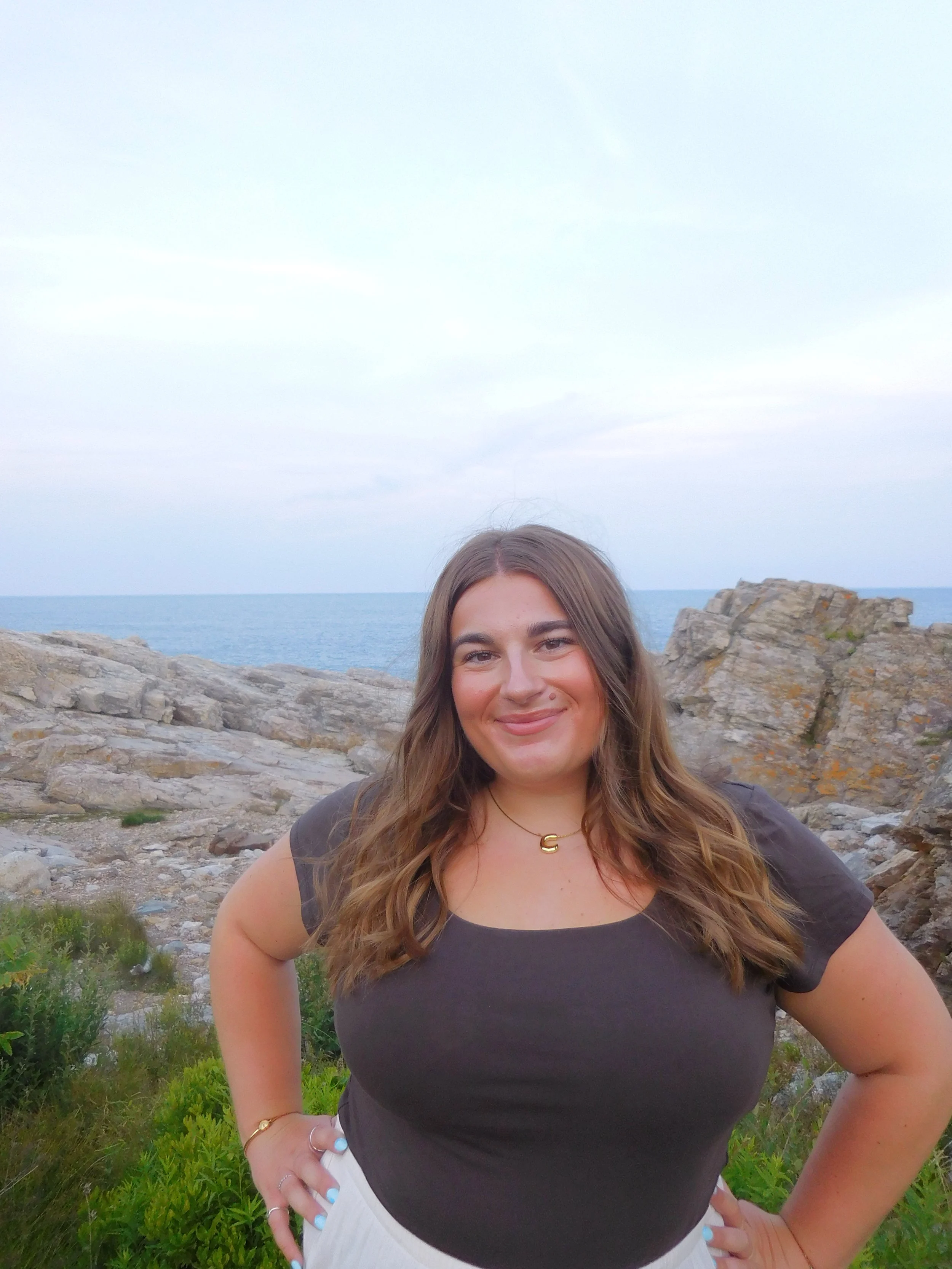 A young woman standing outdoors near rocks and green plants with the ocean and sky in the background, smiling with long brown hair, wearing a dark gray t-shirt, white pants, and jewelry.