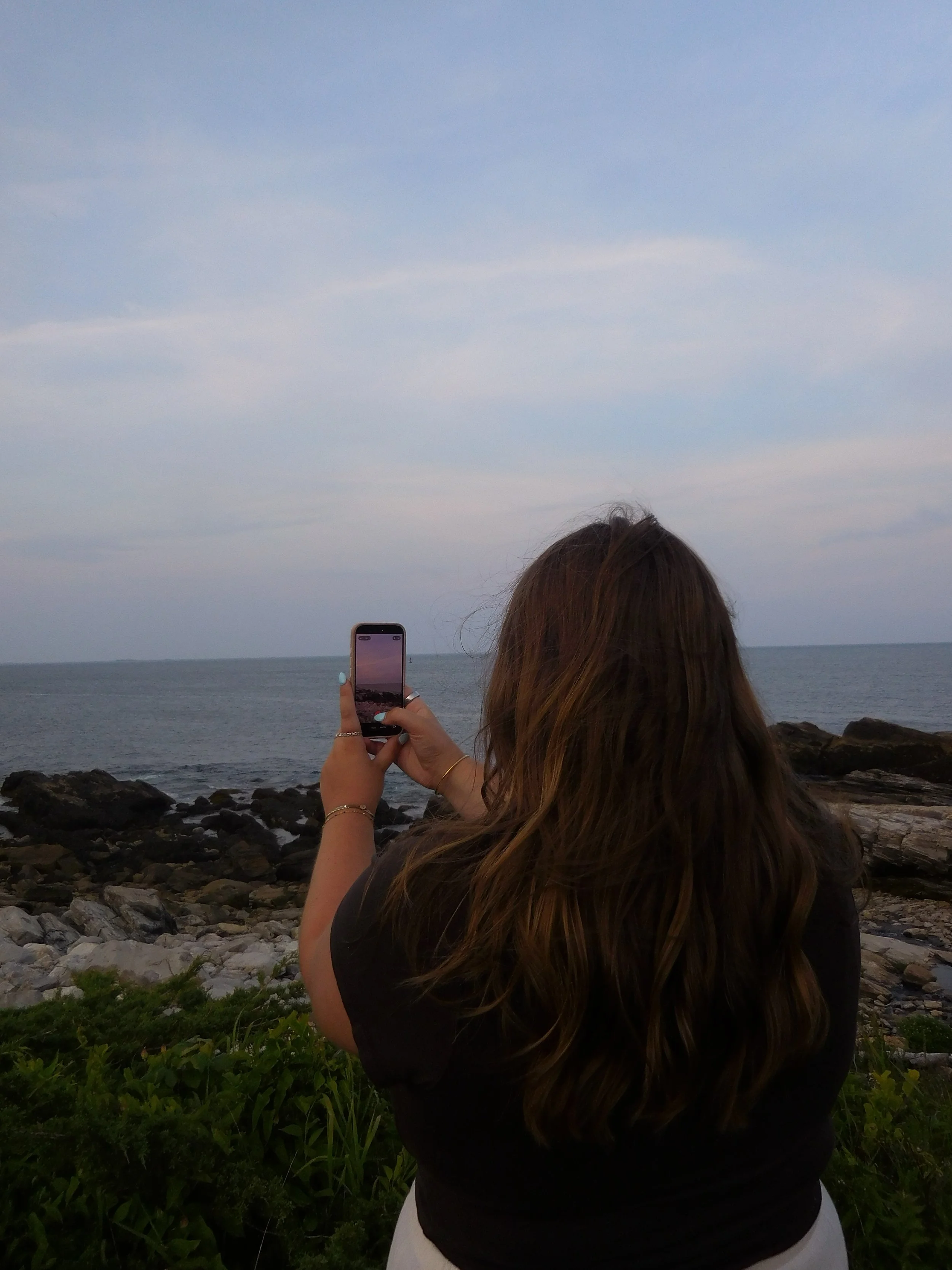A woman with long brown hair taking a photo with her phone of the ocean and sky at sunset, with rocks and greenery in the foreground.
