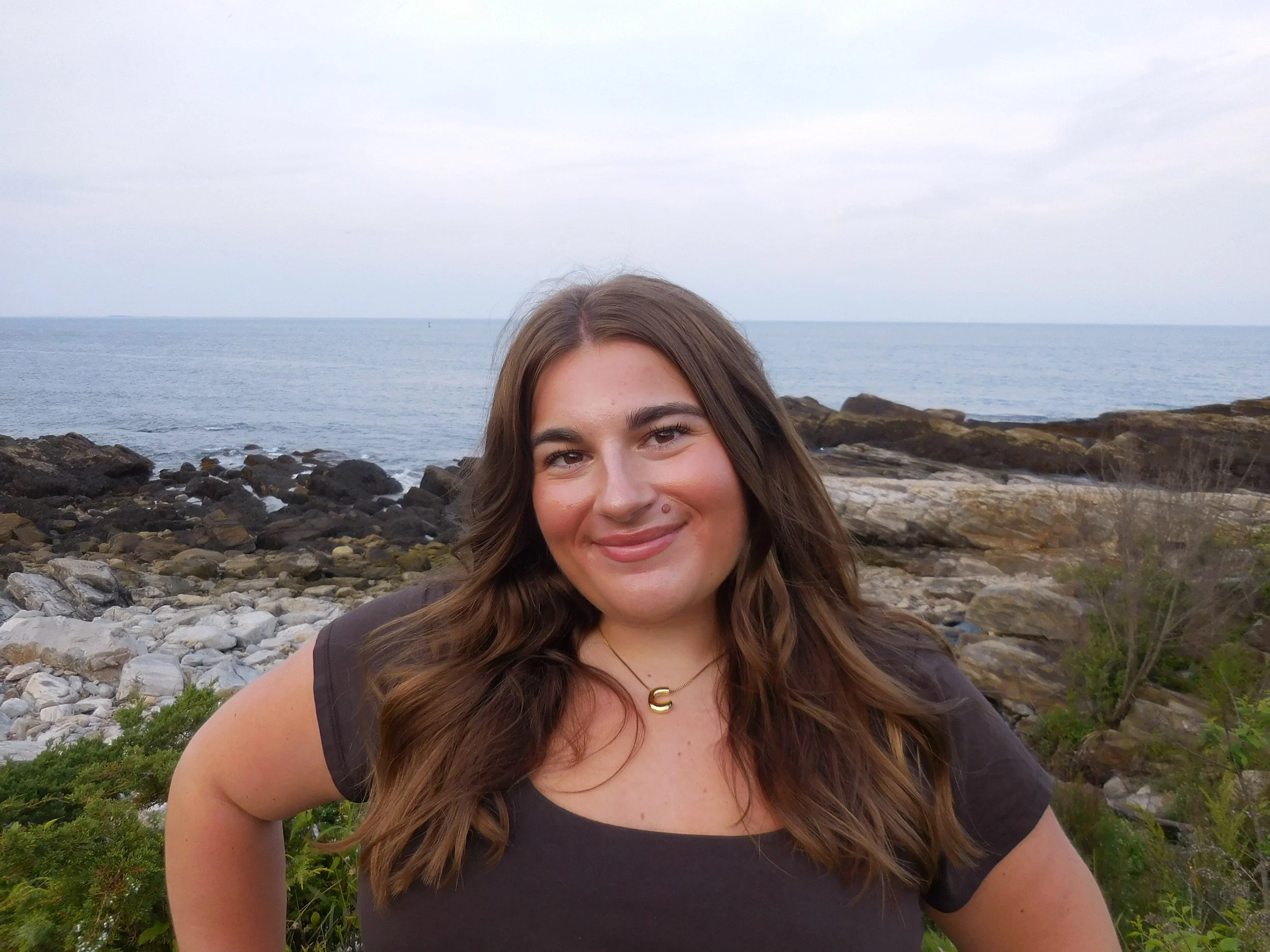 A smiling woman with long brown hair at the rocky shoreline by the ocean, overcast sky in the background.