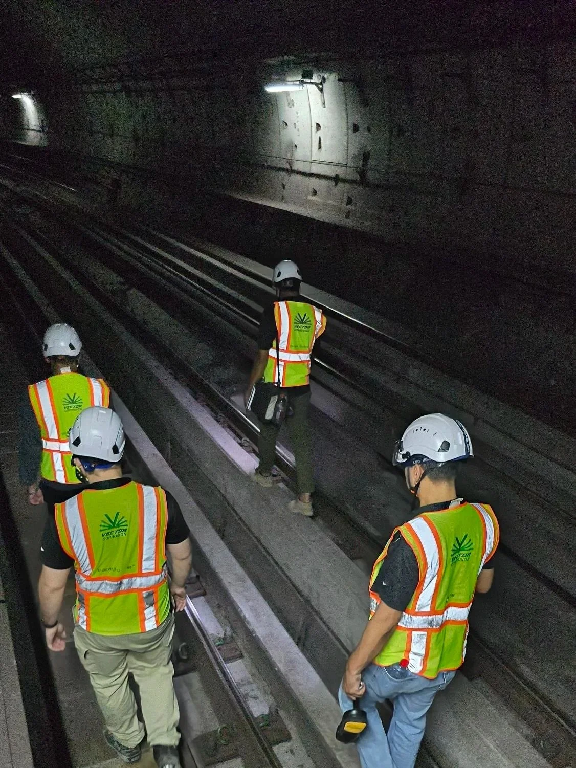 Workers in safety vests and helmets inspecting railway tracks inside a tunnel.