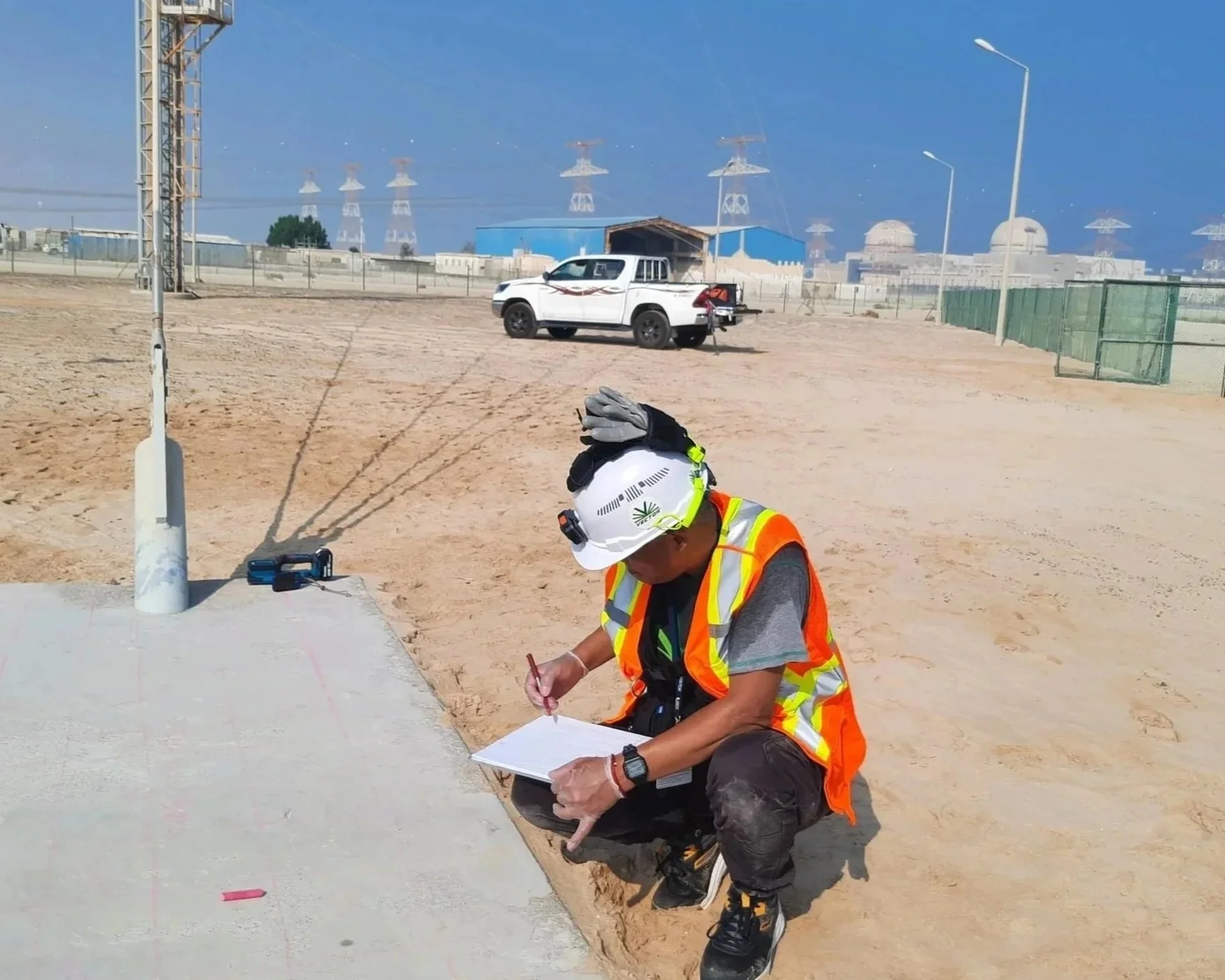 A construction worker wearing a white safety helmet and orange reflective vest is kneeling on the ground, writing or drawing on a surface with a marker. The person is in a sandy, open area with construction equipment and a white pickup truck in the background. There are power lines and industrial buildings in the distance, and the sky is clear and blue.