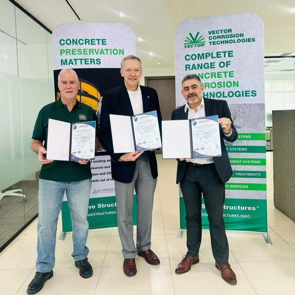 Three employees holding our ISO Certification's stand in front of banners about concrete preservation and corrosion technologies at a professional event.