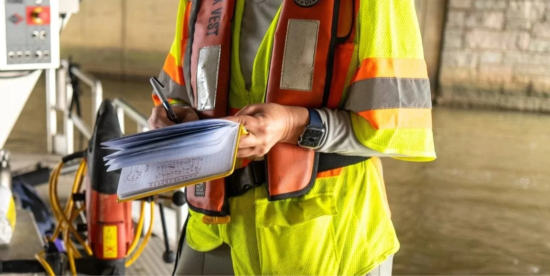 A person wearing a yellow safety vest and a red safety vest with reflective strips, holding a notepad, pen, and a smartwatch, with a wheelbarrow and other equipment in the background, possibly at a construction or industrial site.