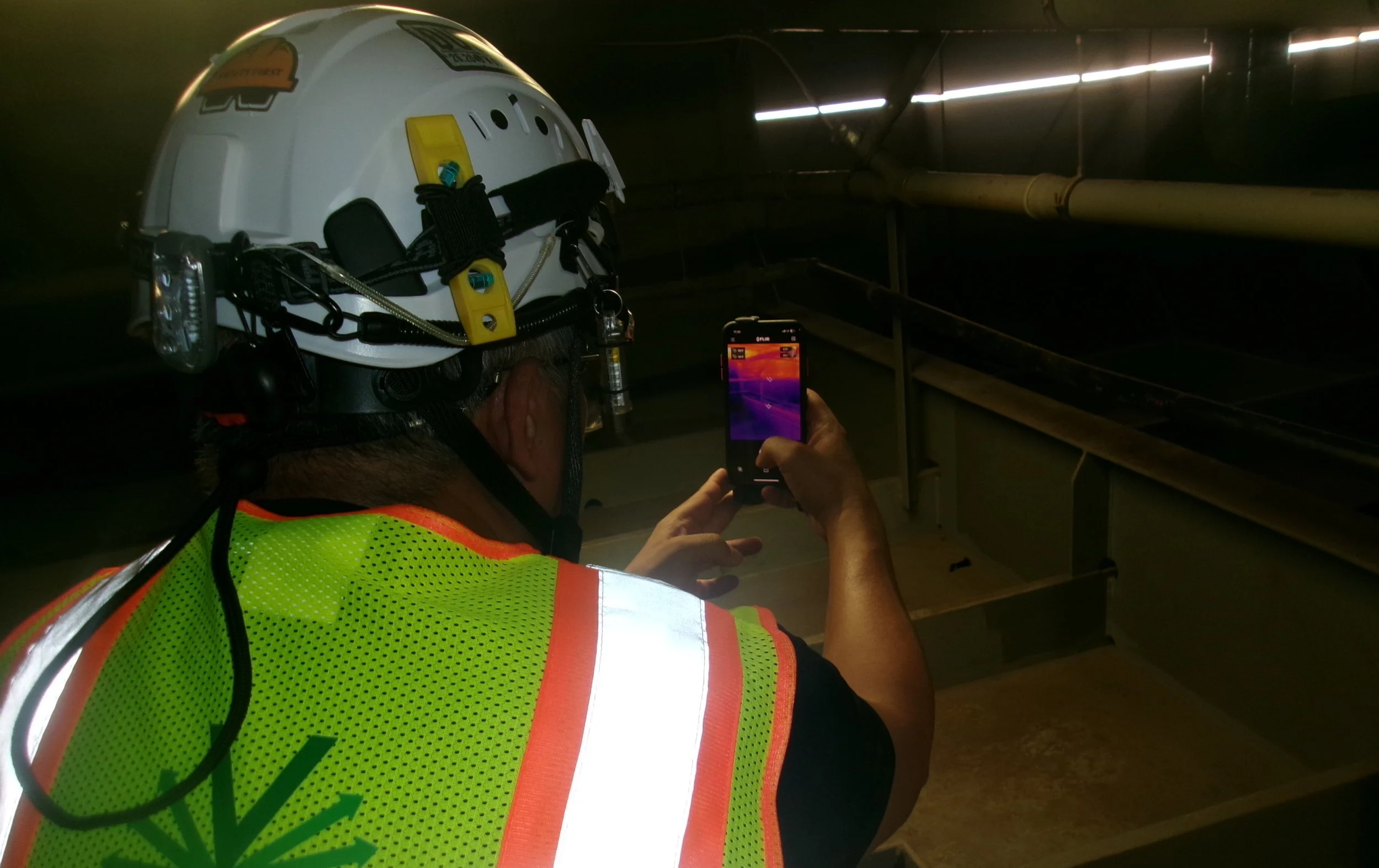 A worker in a neon safety vest and helmet is using a thermal imaging camera to take photos of industrial pipes or ducts in a dark facility.