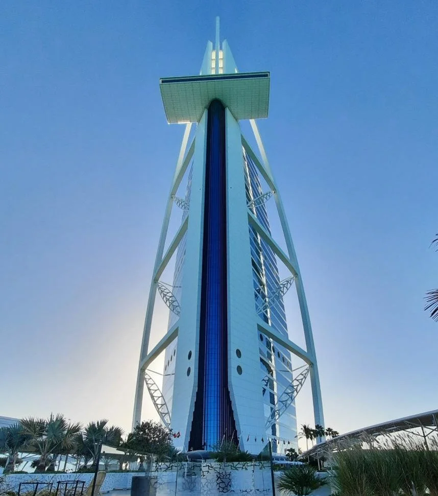 The image shows the Burj Al Arab hotel in Dubai, characterized by its sail-shaped structure and modern architecture, set against a clear blue sky.