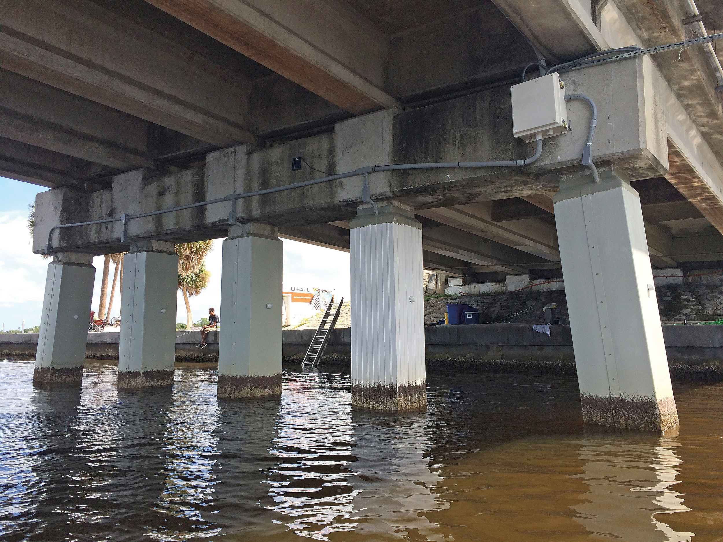 View of the underside of a bridge over water, with galvanic jackets for tidal, splash, underwater and atmospheric zone protection of marine piles.