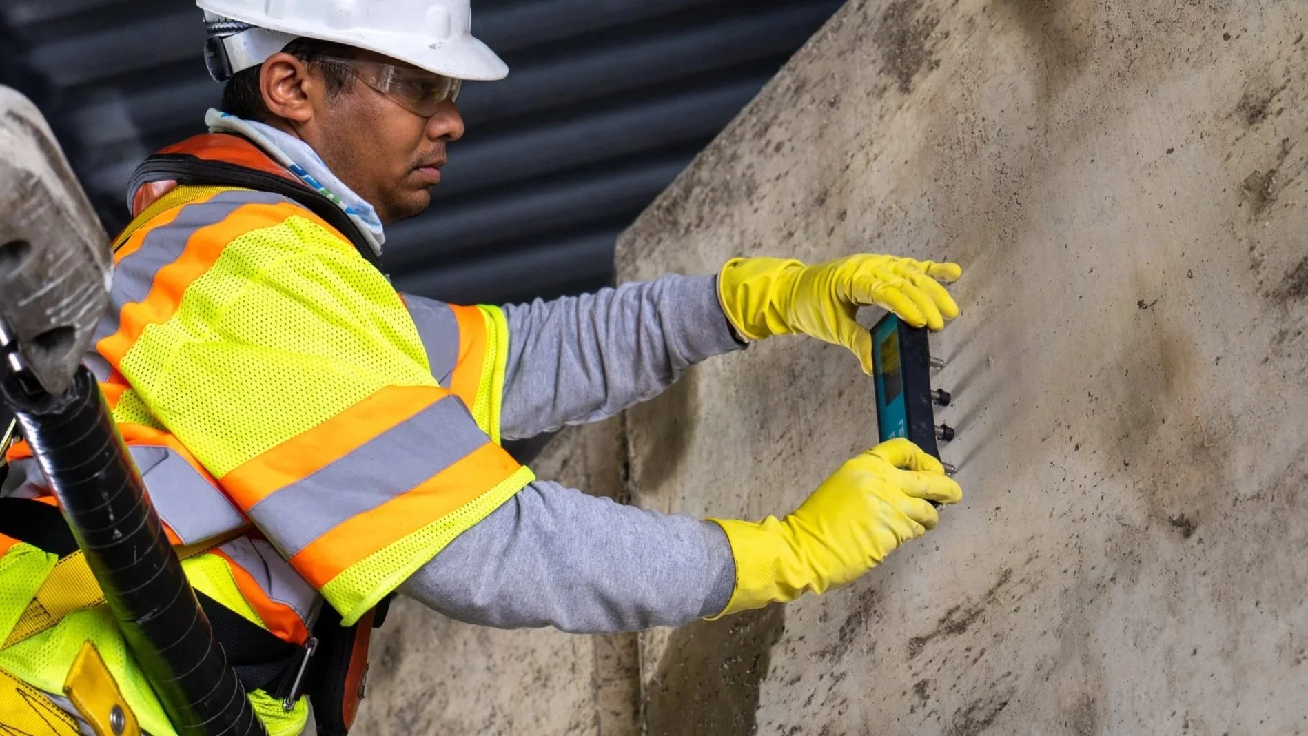 A construction worker wearing a white hard hat, safety glasses, a bright yellow and orange reflective safety vest, gray long-sleeve shirt, and yellow gloves is using a Wenner probe against a large concrete wall.