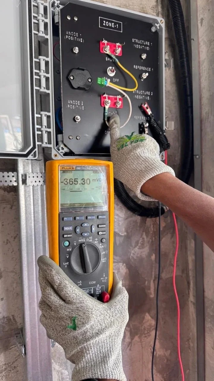 A person wearing gray gloves testing a monitoring box with a digital multimeter, reading -365.30 millivolts.