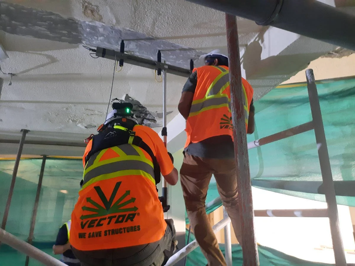 Two construction workers in orange safety vests using an impact echo/pulse velocity device on building a ceiling or wall, surrounded by scaffolding and green safety barriers.