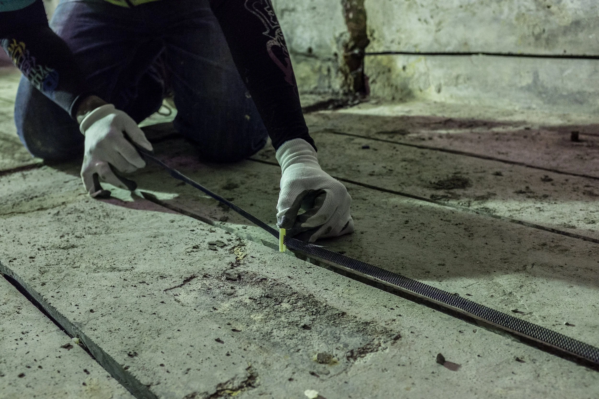 Person kneeling on the ground in a construction or renovation site, installing or a cathodic protection design on concrete surface. The person is wearing white gloves and dark clothing.