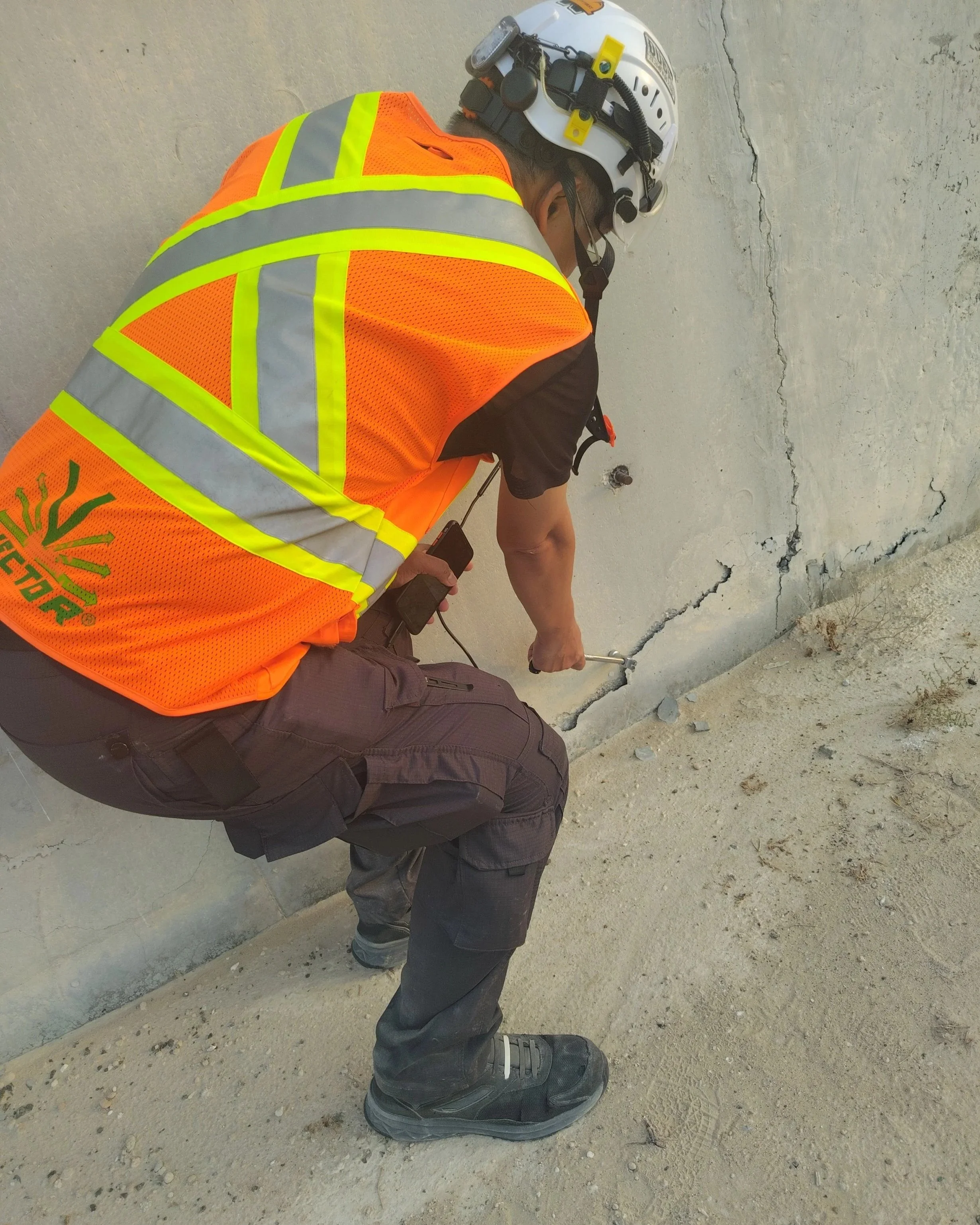 A construction worker wearing a white helmet, orange safety vest with reflective stripes, black cargo pants, and black work boots is examining a cracked concrete wall with a crack running vertically. The worker is kneeling on the ground with one hand holding a tool, possibly inspecting or measuring the crack.