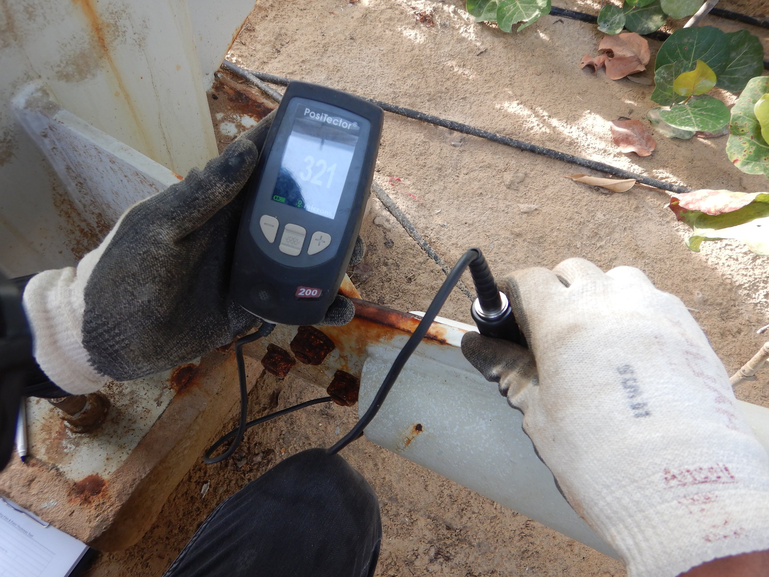 A hand holding a ultrasonic thickness (UT) gauge against structural steel supports and baseplates.