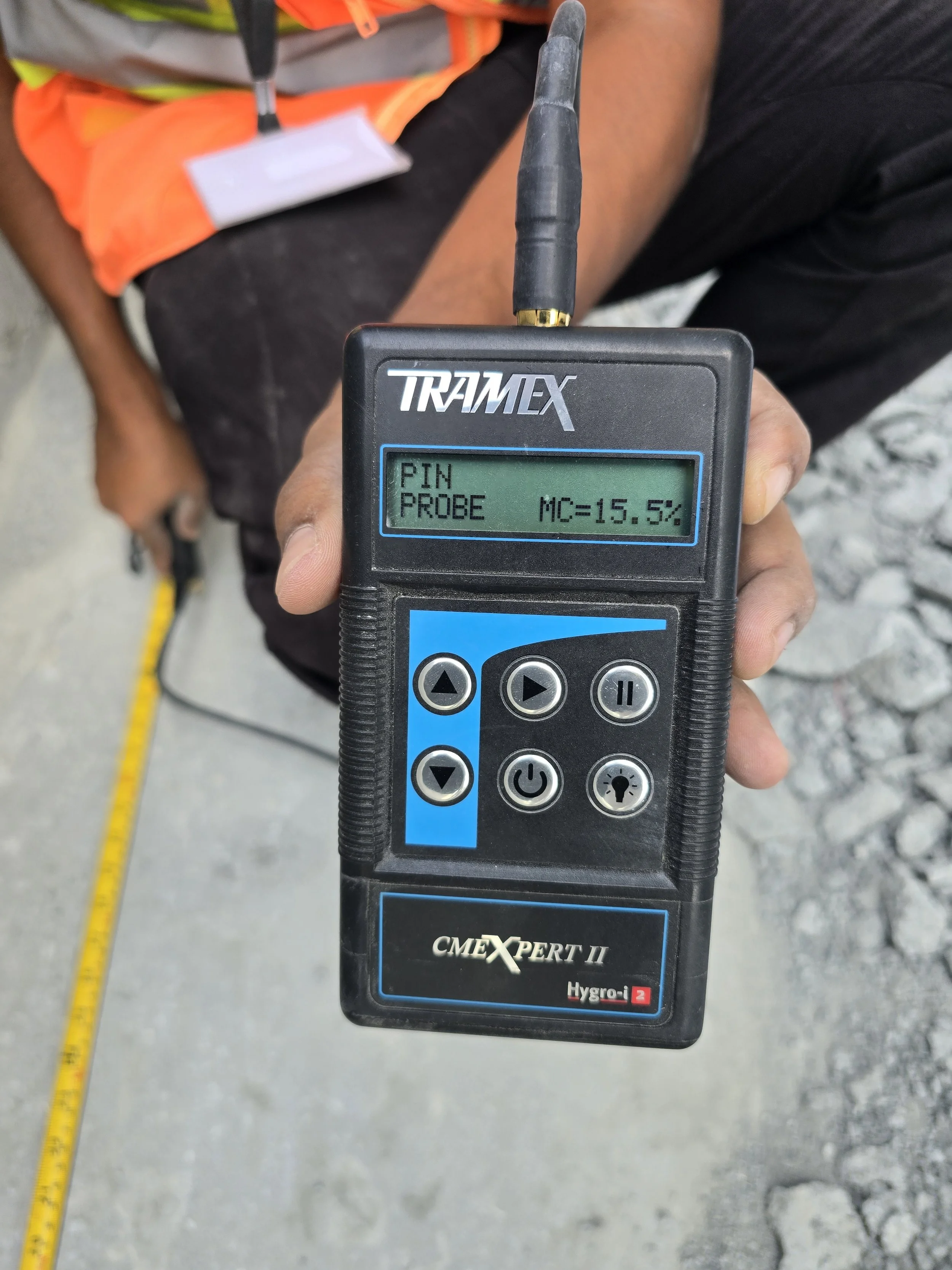 A moisture meter displaying a reading of 15.5% with the label 'PIN PROBE' above it. In the background, a person is wearing a high-visibility safety vest and holding the device, with a tape measure and moisture meter probe on a concrete surface.