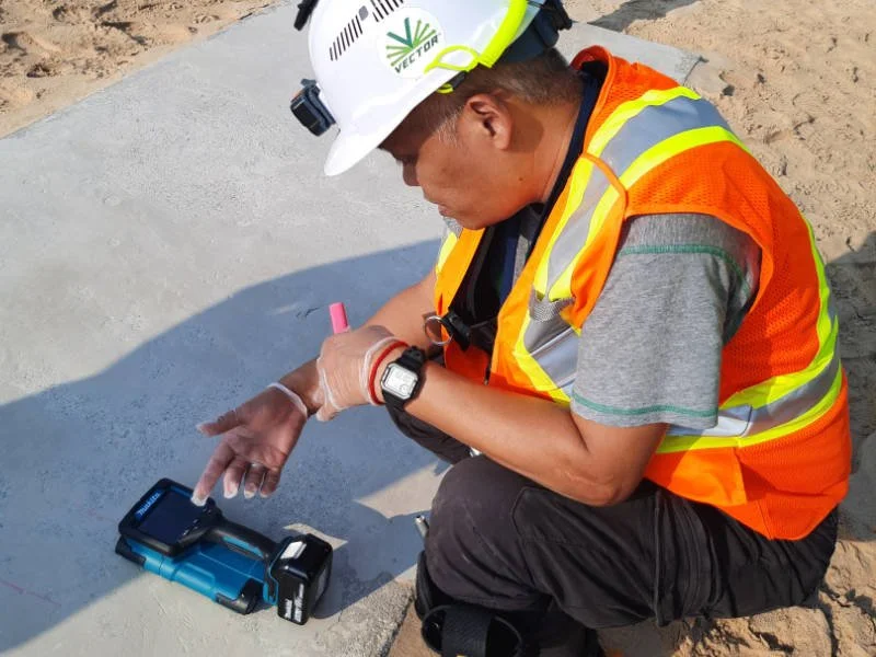A construction worker wearing a white safety helmet, orange safety vest, gray t-shirt, and watch kneels on the ground, operating a handheld ground penetrating radar device placed on a concrete surface at a construction site.