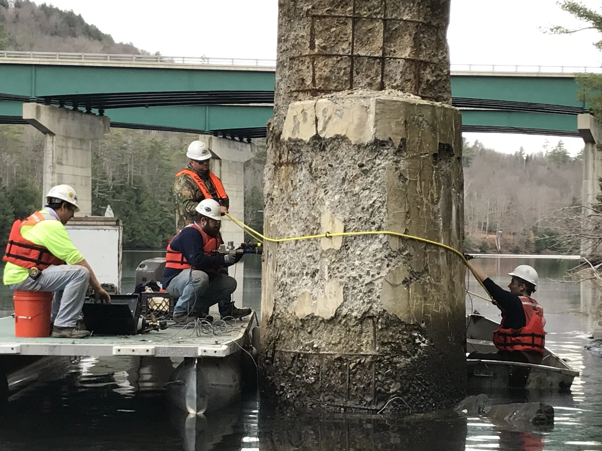 VCME workers on a pontoon barge conducting an acoustic tomography test with a sensor chain wrapped around a submerged bridge pile, wearing full PPE.